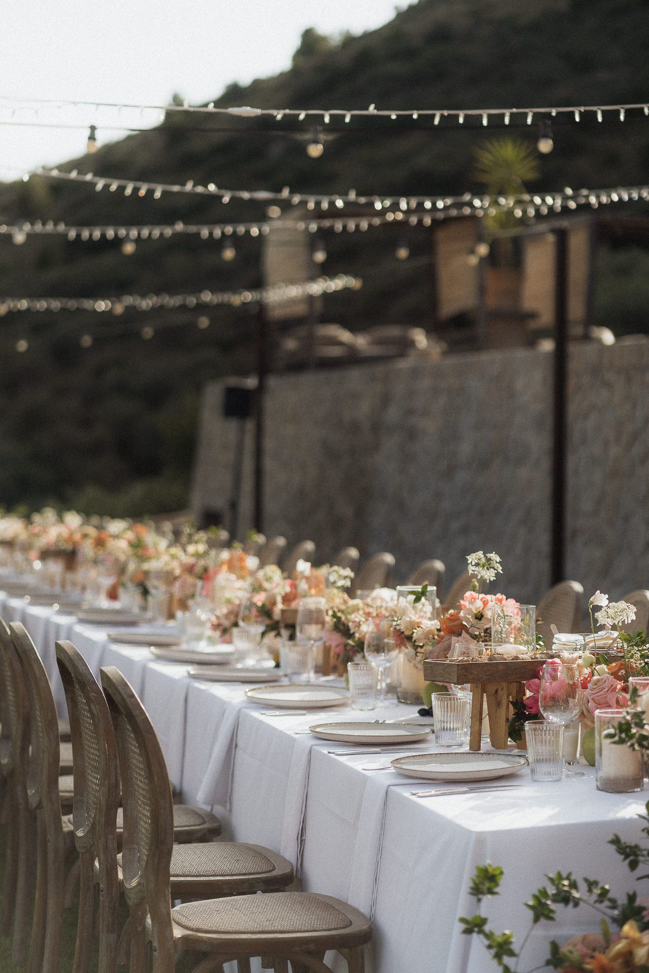 Elegant outdoor wedding reception table with white linen, pink floral centerpieces, and string lights overhead.