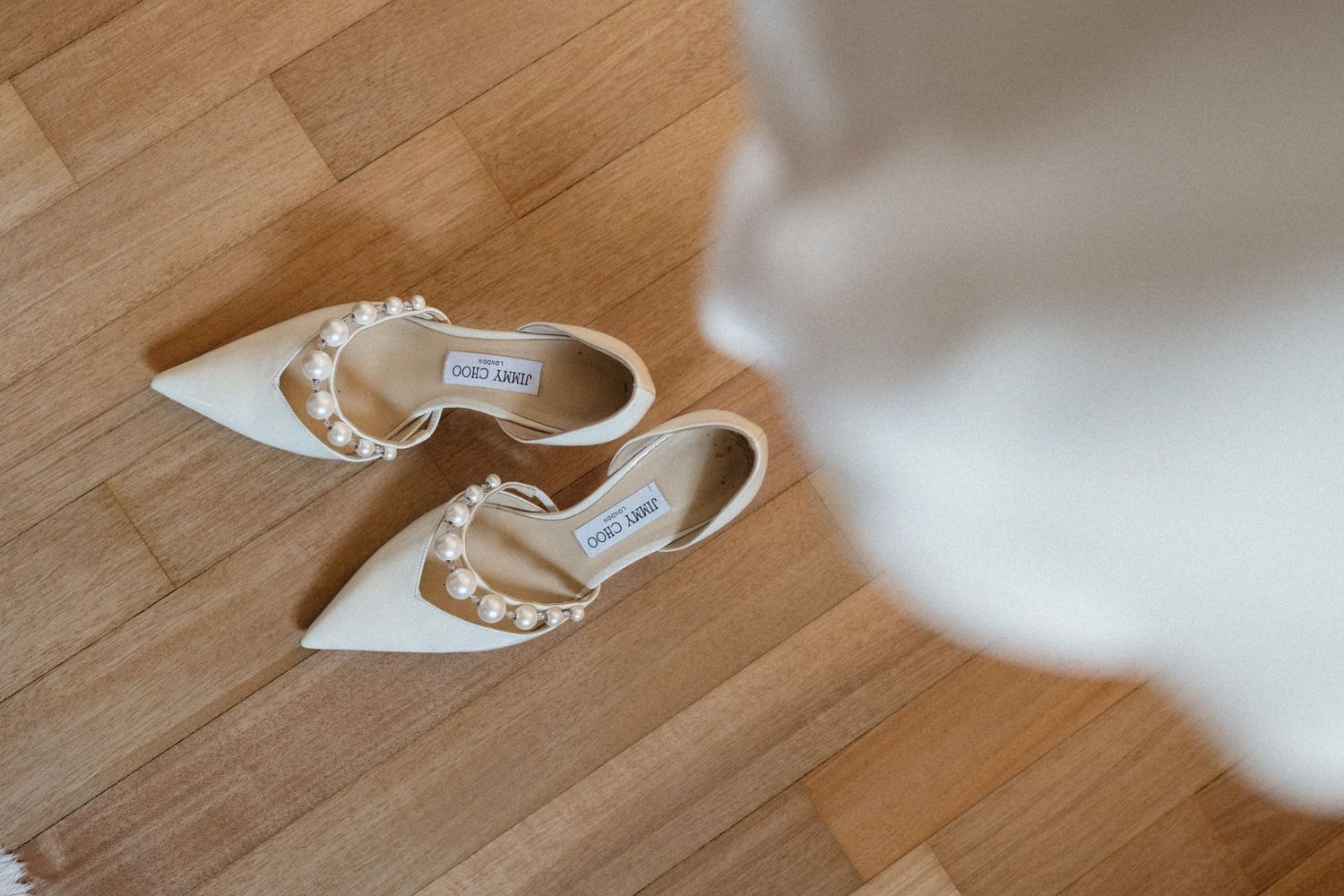 Elegant white Jimmy Choo bridal heels with pearl embellishments displayed on wooden floor with wedding dress.