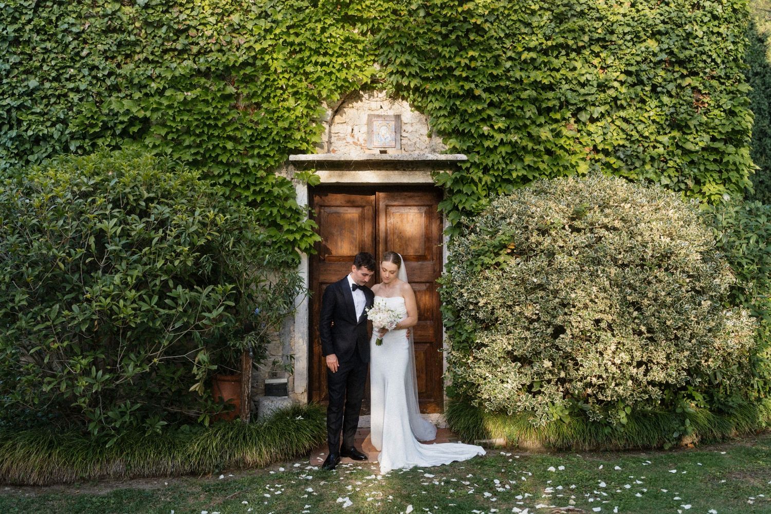 Bride and groom embracing before an ivy-covered stone doorway in a lush garden setting.