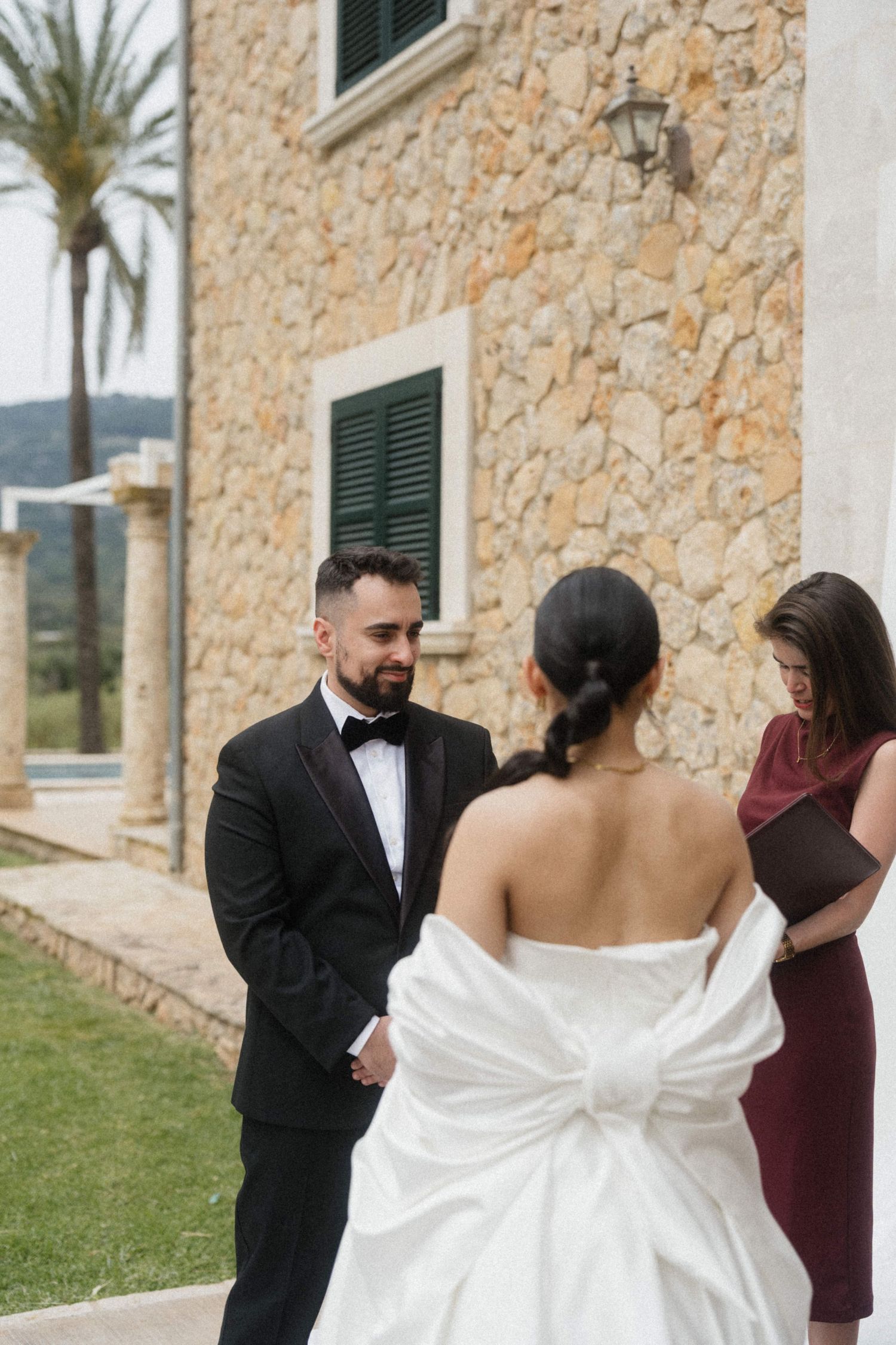 Groom in black tuxedo smiling at bride in white strapless wedding dress during outdoor ceremony.