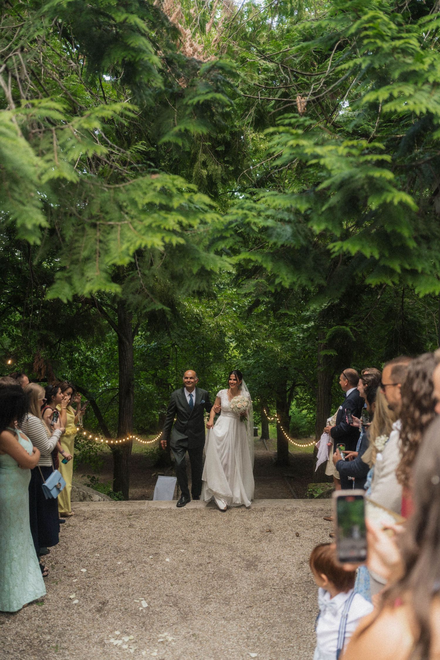 Bride and groom walking down outdoor aisle lined with guests and string lights among trees.