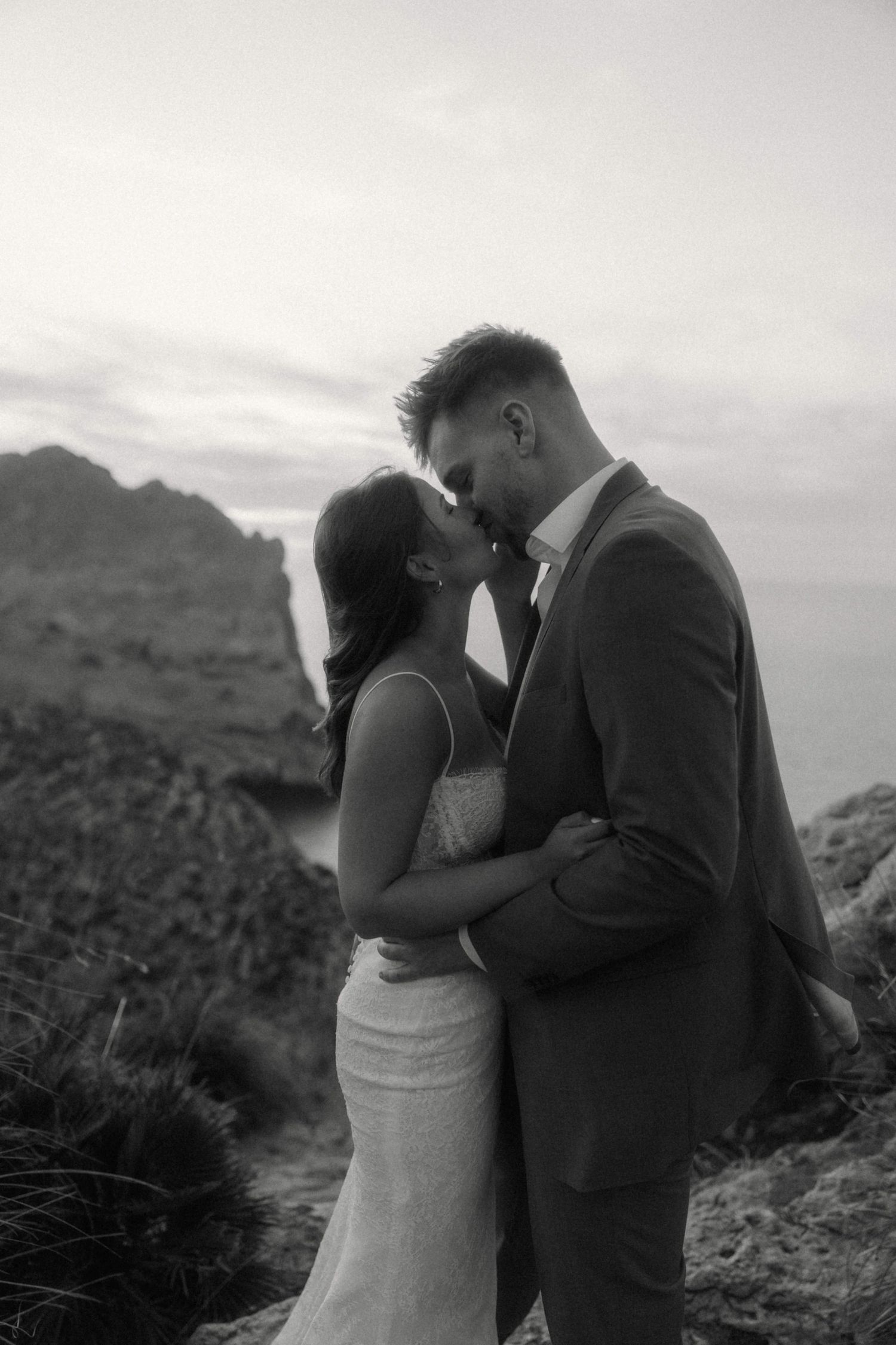 Black and white photo of bride and groom sharing a kiss on rocky coastal cliffside at dusk.