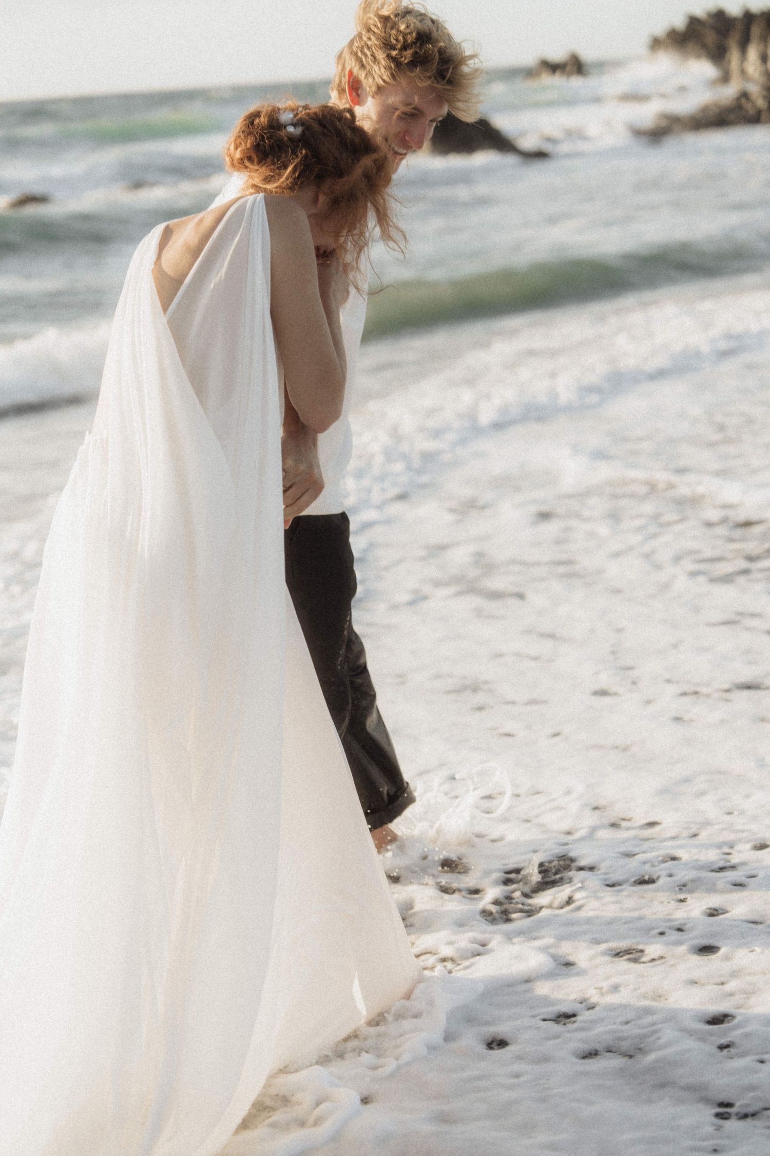 Bride in flowing white gown embracing groom at water's edge with ocean waves at beach elopement.