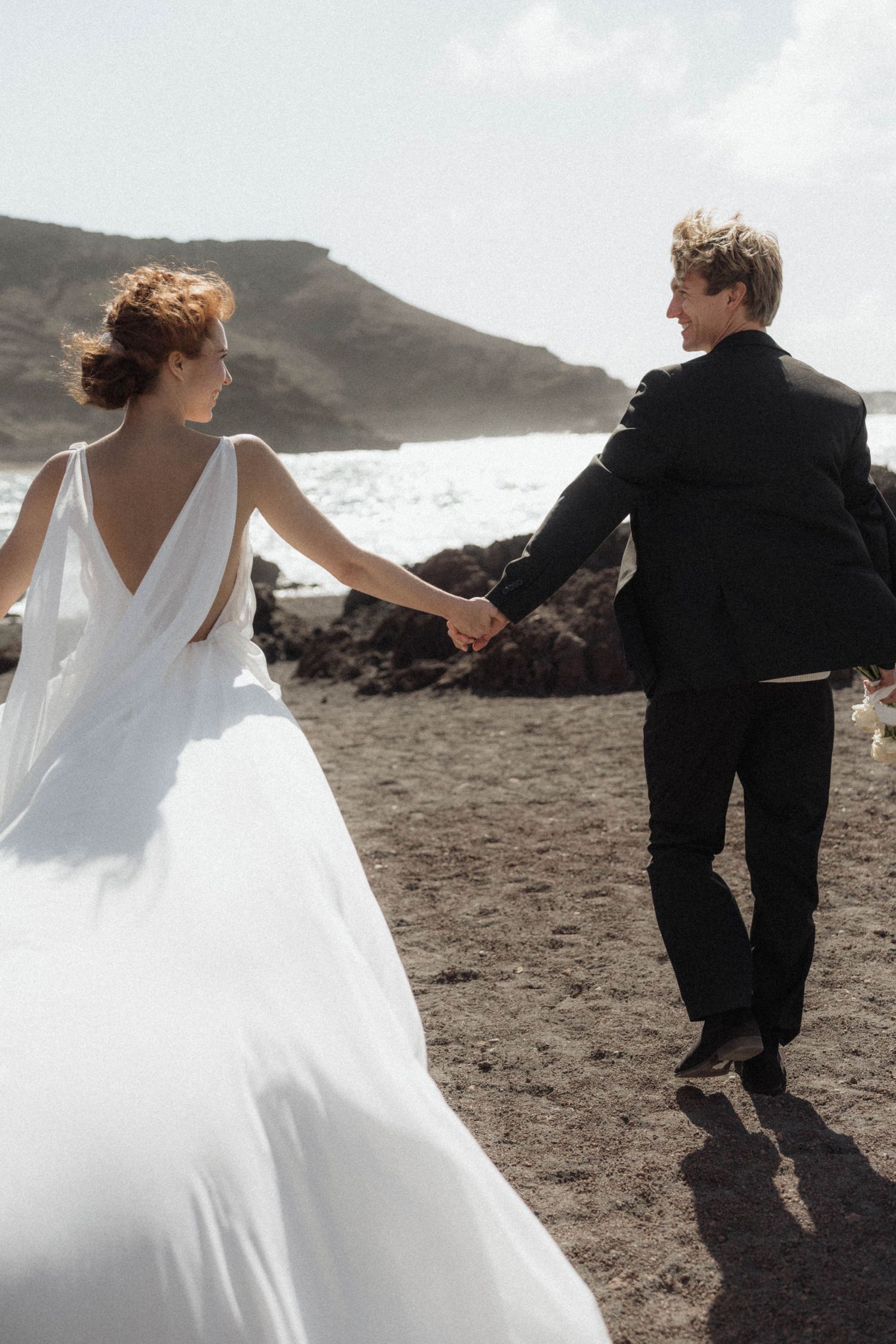 Bride and groom hold hands walking on dark sandy beach with ocean waves behind them.