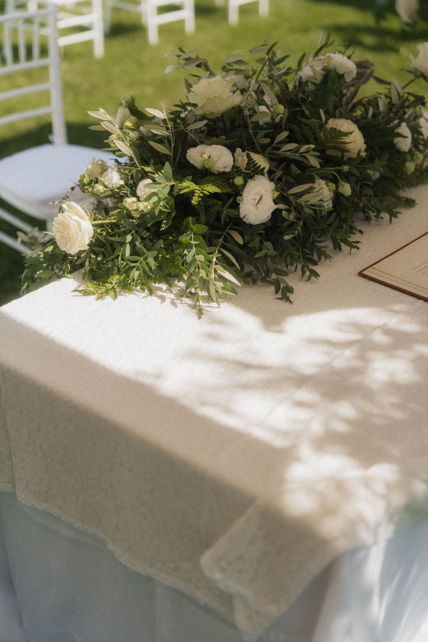 Elegant outdoor wedding table with white linen and floral arrangement of white roses and greenery in sunlight.