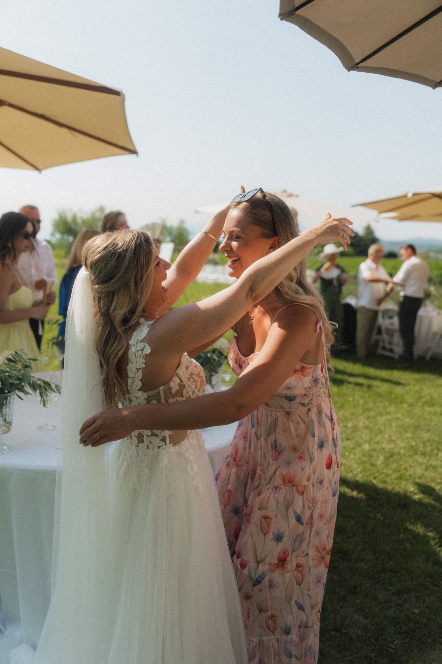 Bride in white gown embracing smiling guest in floral dress at outdoor wedding.