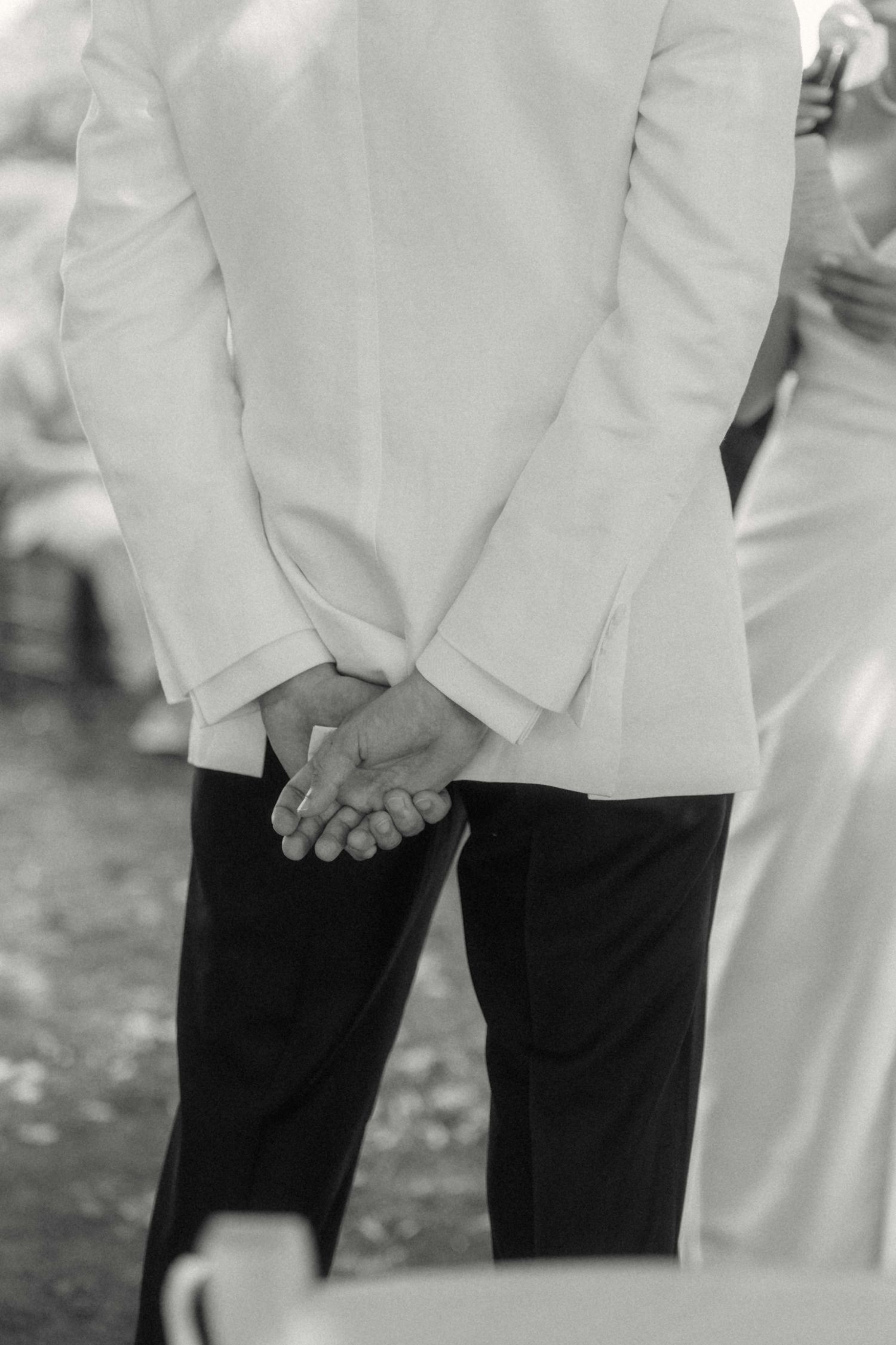 Black and white close-up of groom in white jacket clasping hands behind his back at wedding ceremony.