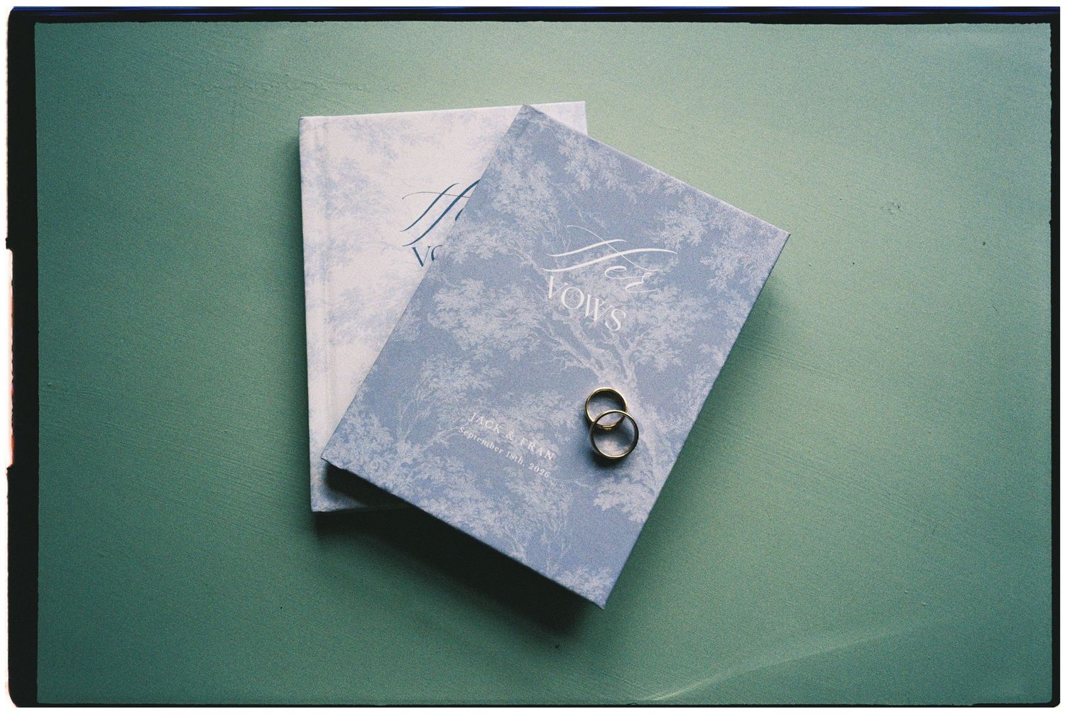 Two blue and white marbled wedding vow books with two gold wedding rings resting on top, set against a soft green background.