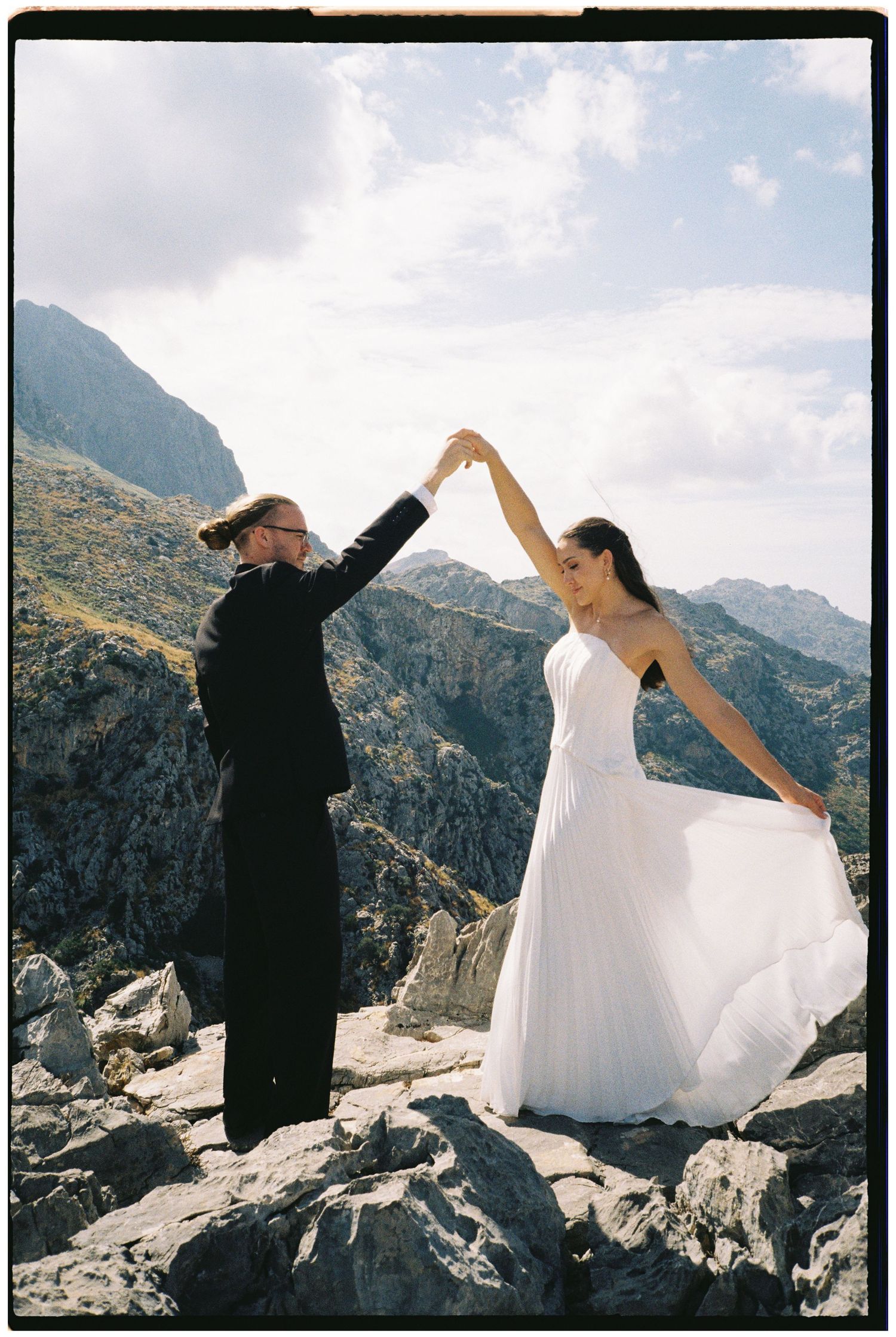 Bride in a flowing white gown and groom in a black suit hold hands and twirl on a rocky mountain summit under a bright, cloudy sky.
