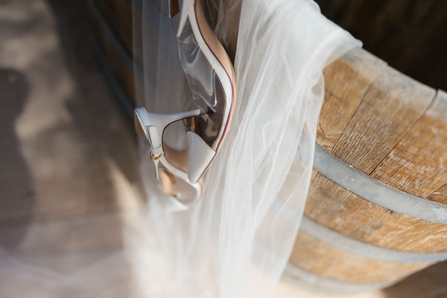 Sheer white tulle veil draped over a rustic wooden barrel beside a pair of blush strappy bridal heels on a sunlit floor.