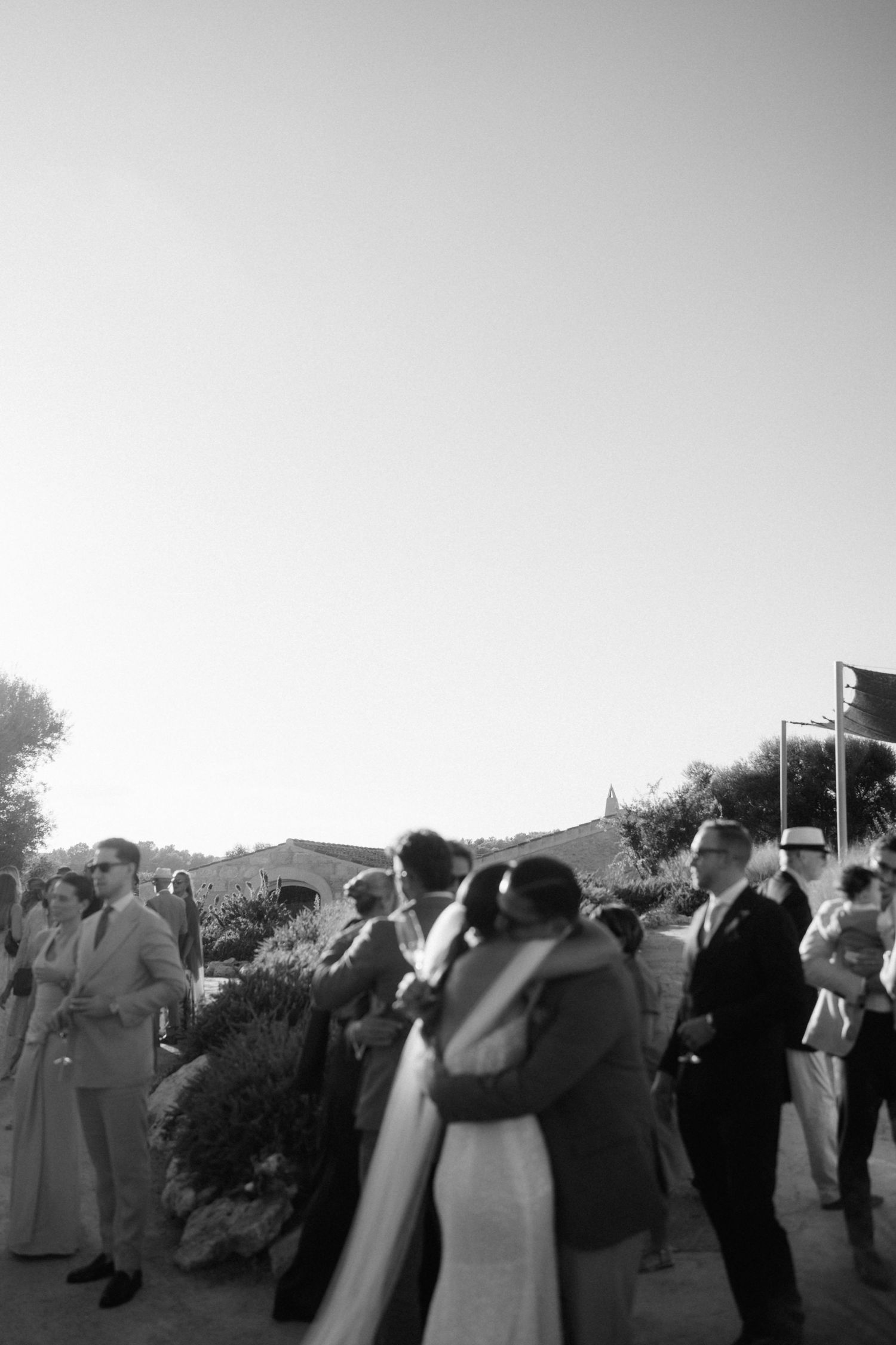 Black and white photo of the bride and groom embracing a guest while a crowd gathers outdoors at a wedding.