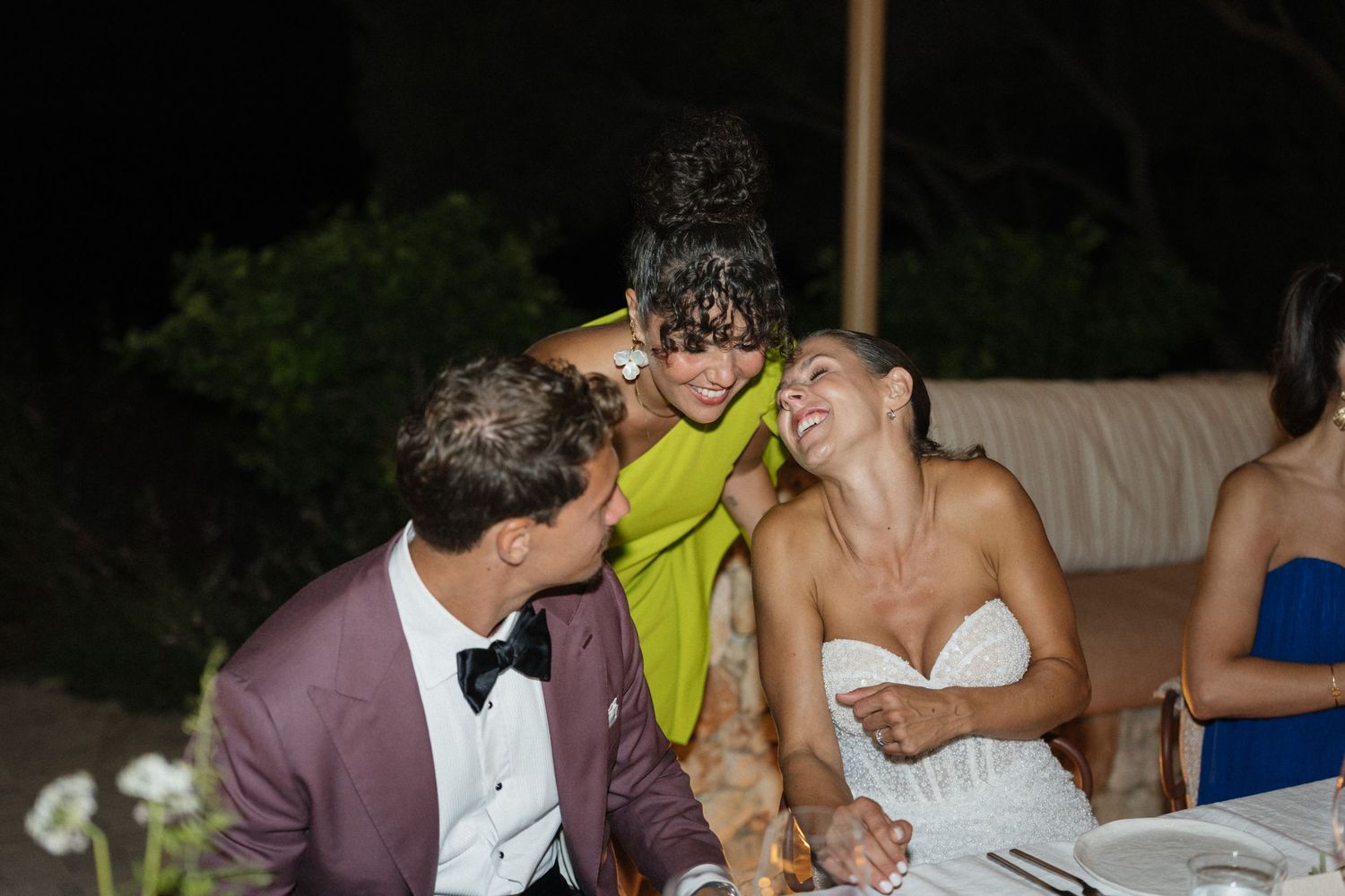 A bride laughs joyfully with a groom in a burgundy blazer and a woman in a green dress at a candlelit evening wedding reception.