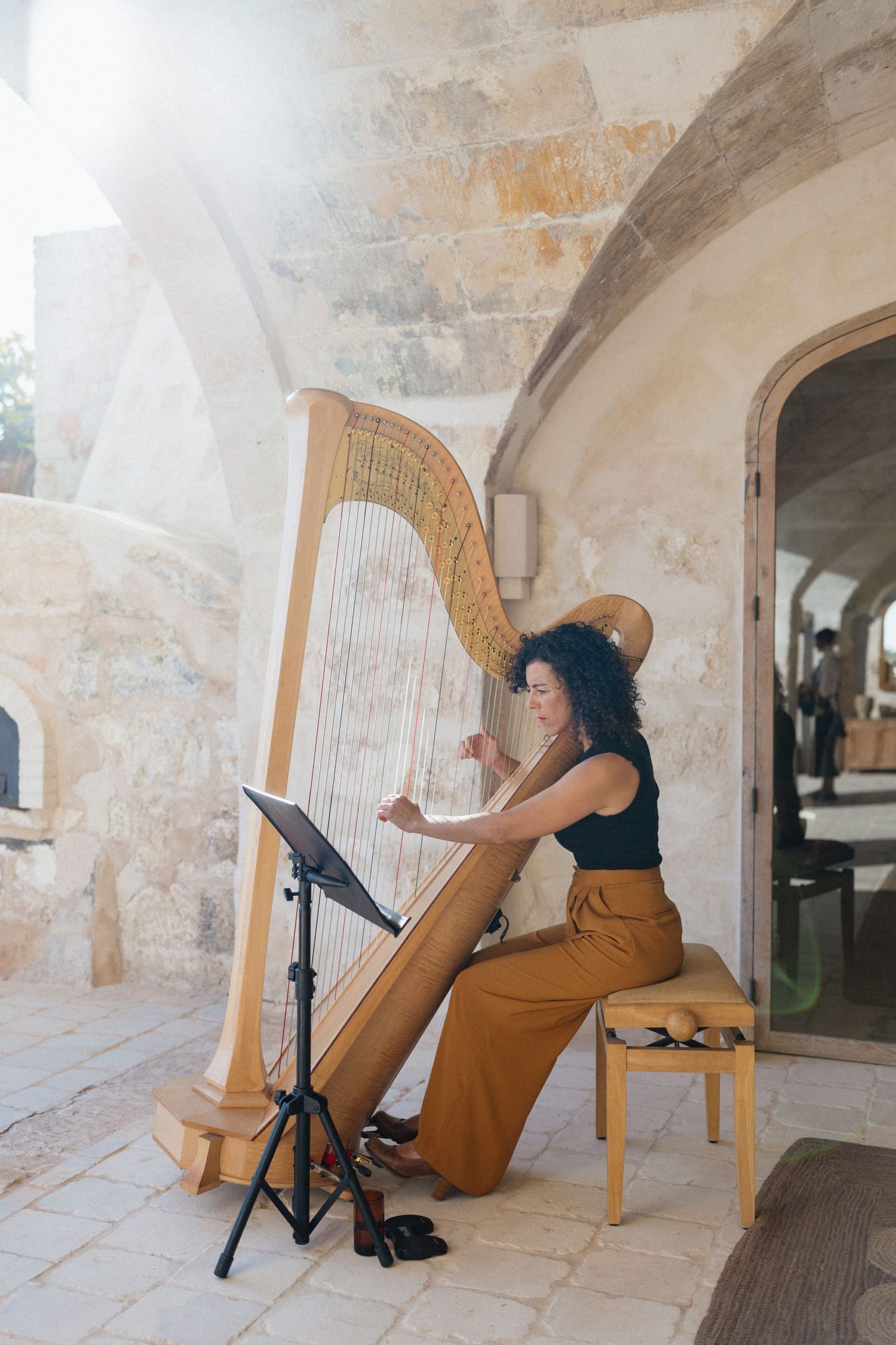 A woman with curly dark hair plays a large harp on a sunlit stone terrace at a Mediterranean wedding venue.