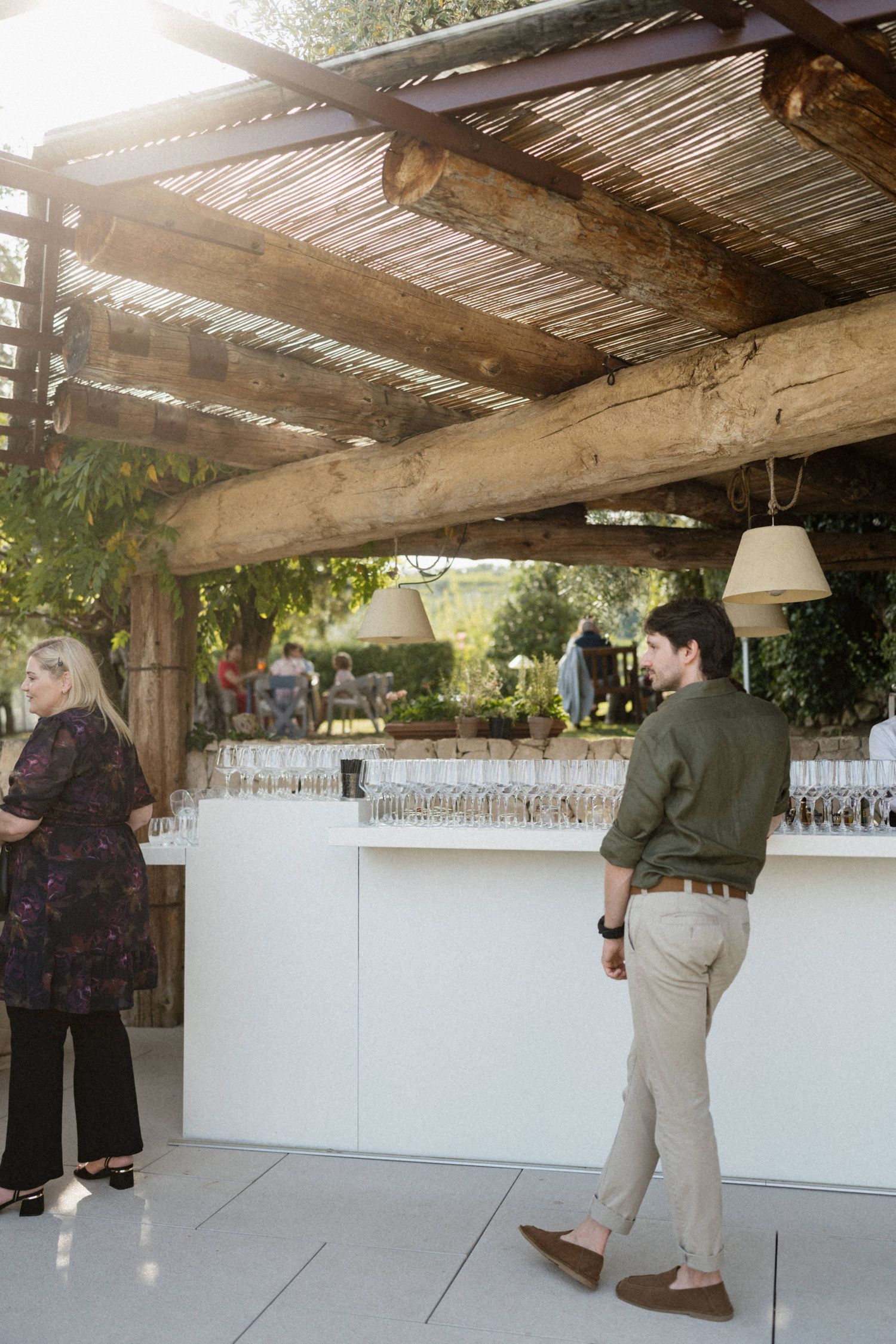 Guests mingle at an outdoor bar under a rustic wooden pergola at an elegant wedding reception venue.