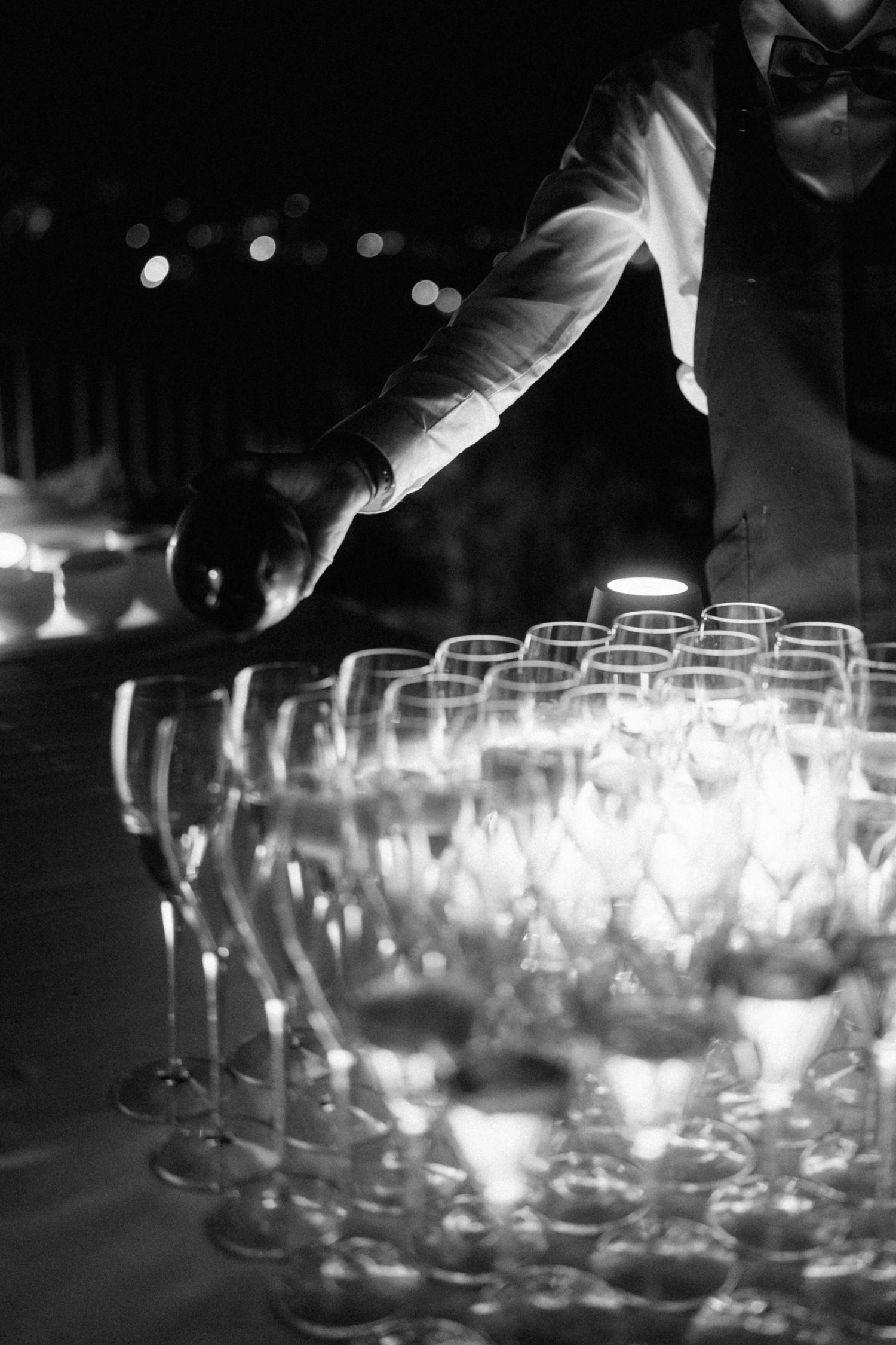 Black and white photo of waiter pouring wine into rows of elegant crystal glasses at night event.