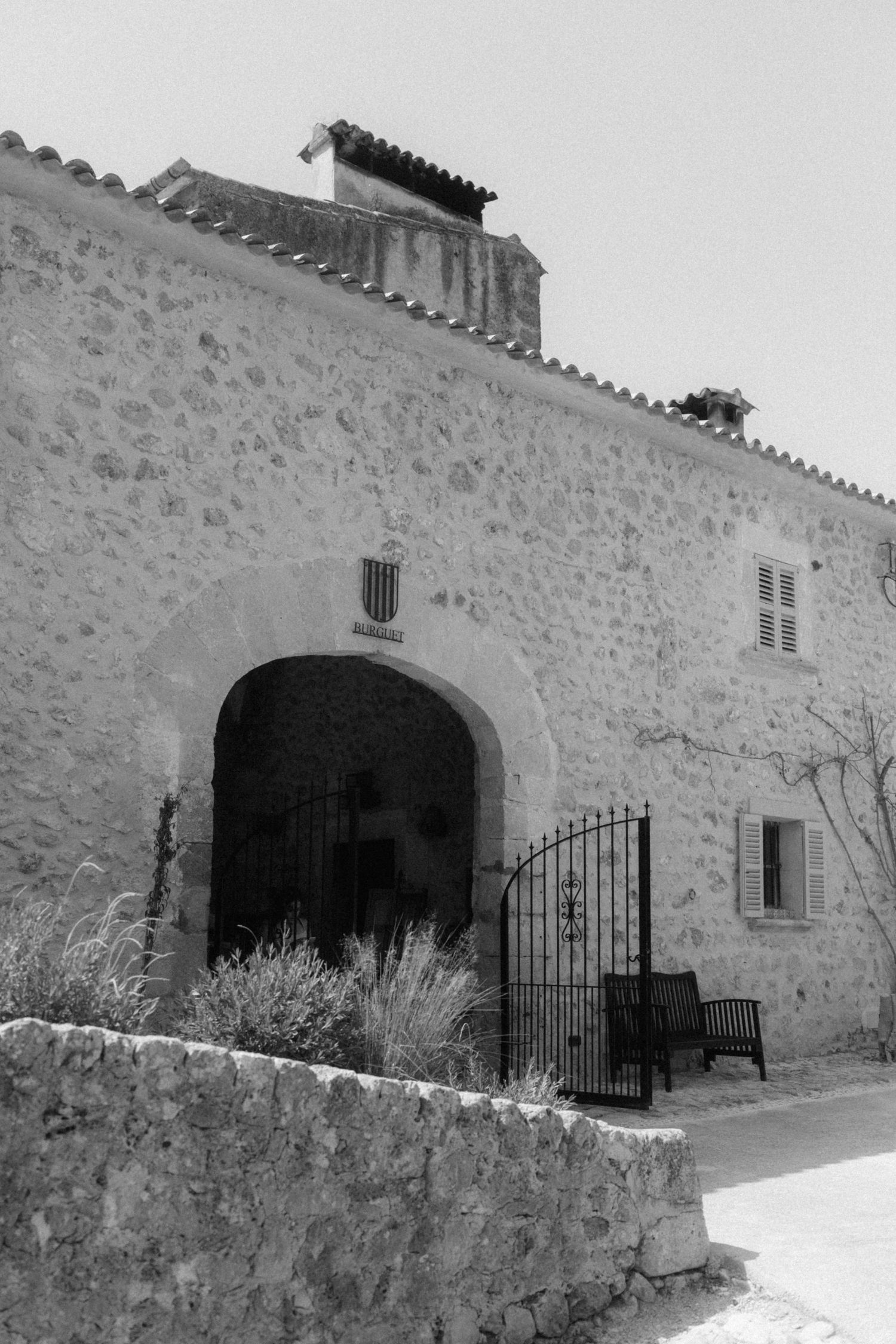 Black and white photo of a rustic stone building with an arched entrance gate and wrought iron fence in Mallorca.