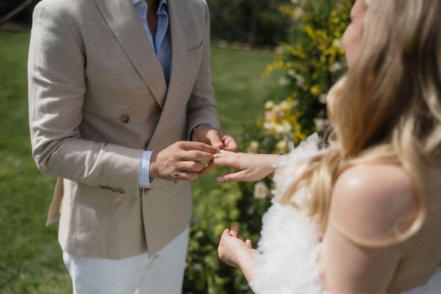 Groom in a beige suit placing a wedding ring on the brides finger during an outdoor ceremony.