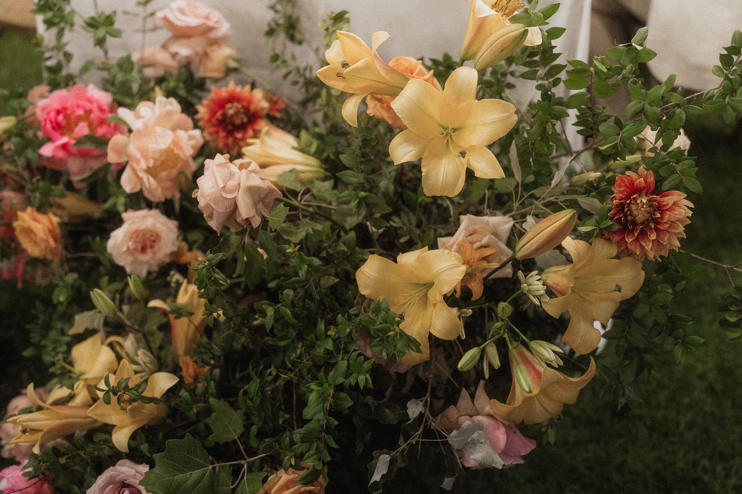 Lush floral arrangement featuring yellow lilies, pink roses, and coral dahlias with green foliage at an outdoor event.