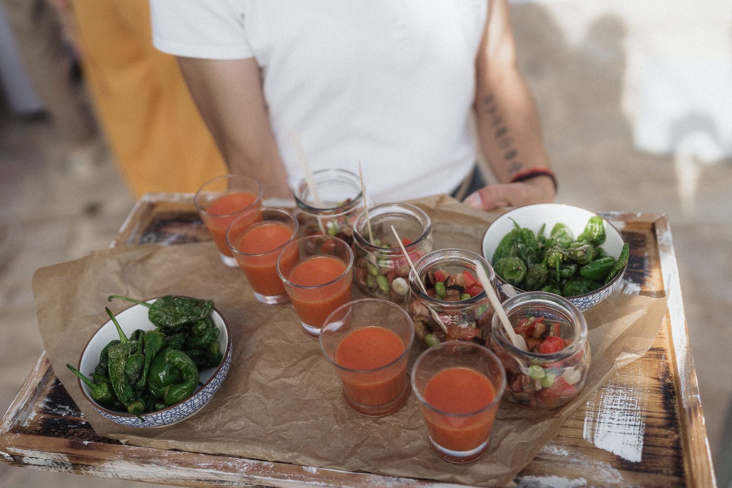 Person holding a wooden tray with Bloody Mary cocktails, fresh vegetables, and appetizers for outdoor entertaining.