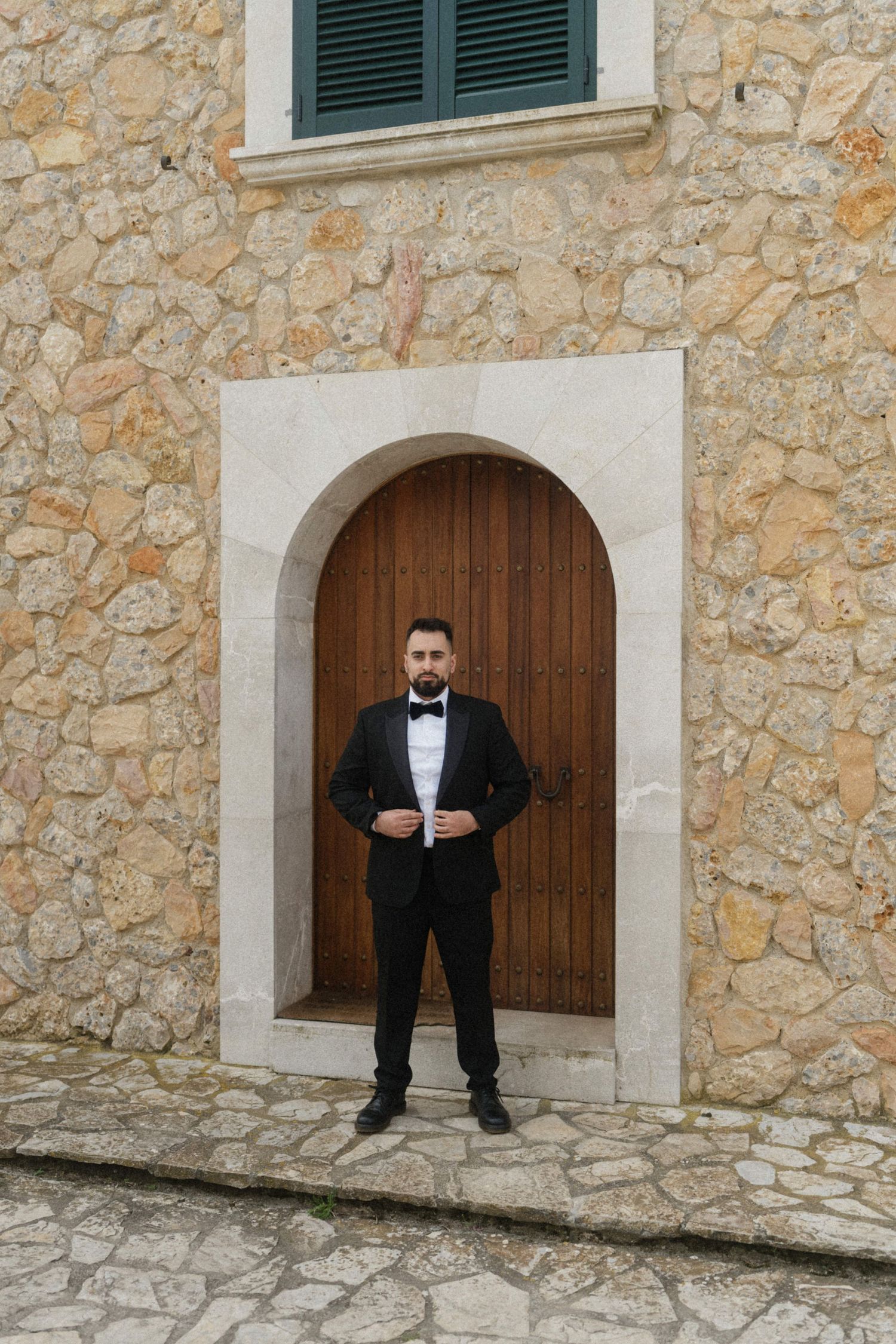 Groom in black tuxedo and bow tie standing before arched wooden door in stone building.