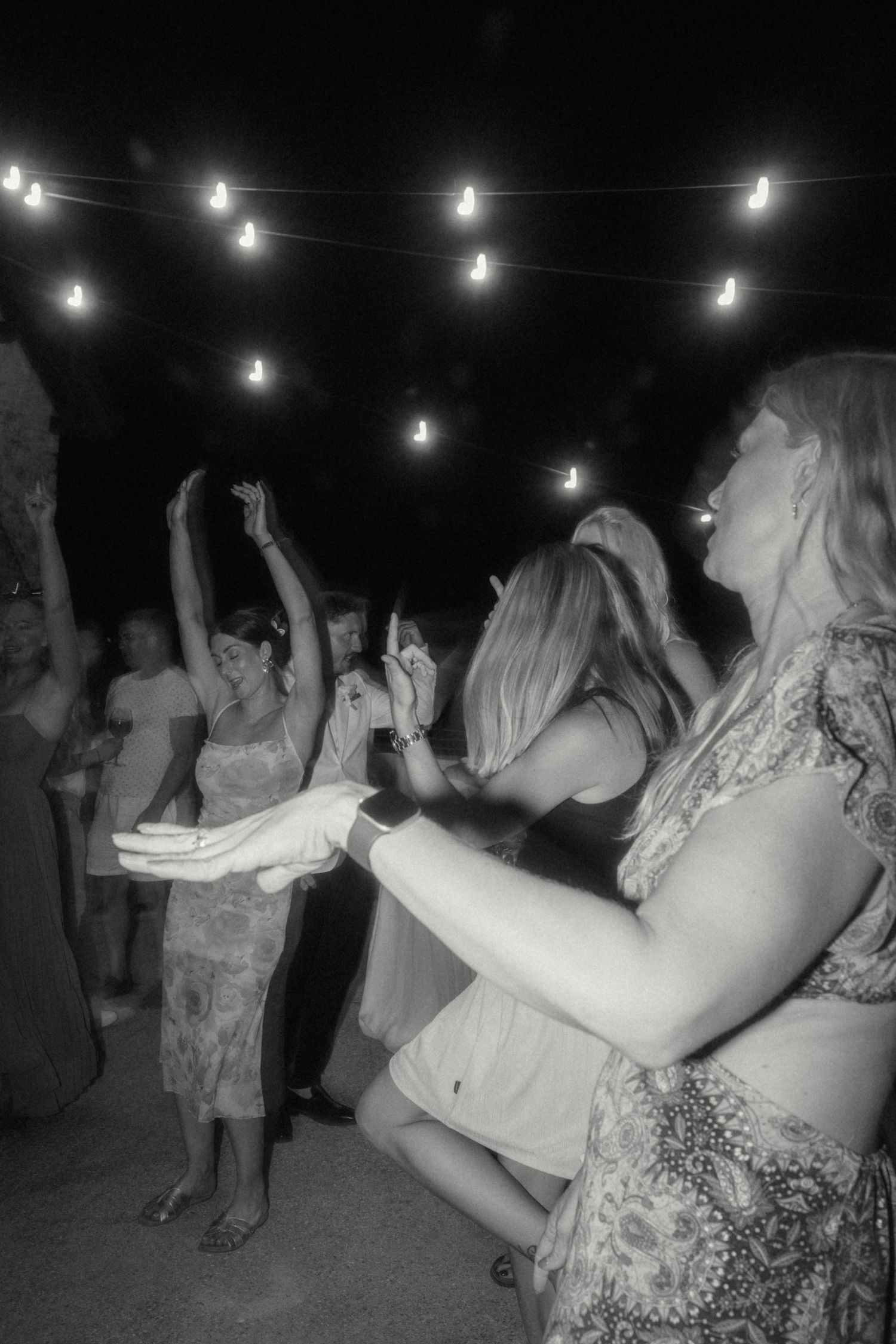 Black and white photo of wedding guests dancing under string lights at night.