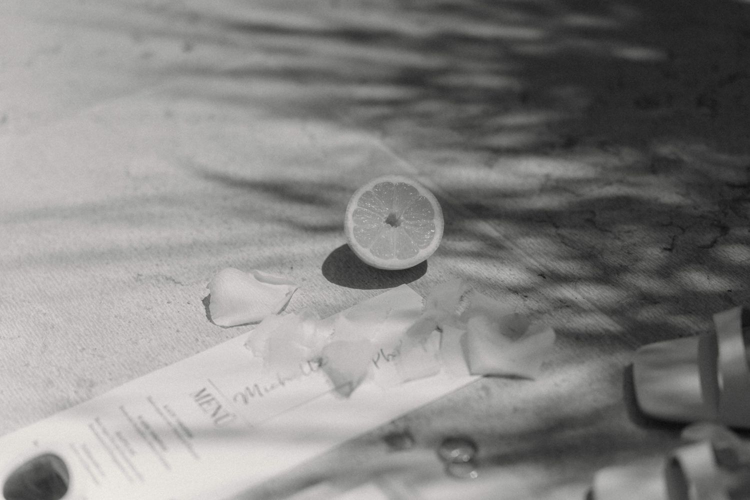 Black and white photo of a halved lemon and ice cubes on a sandy surface with dappled light shadows.