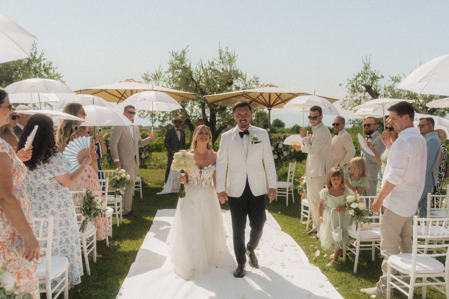 Bride and groom walk down the aisle at an outdoor wedding ceremony with white umbrellas and guests.