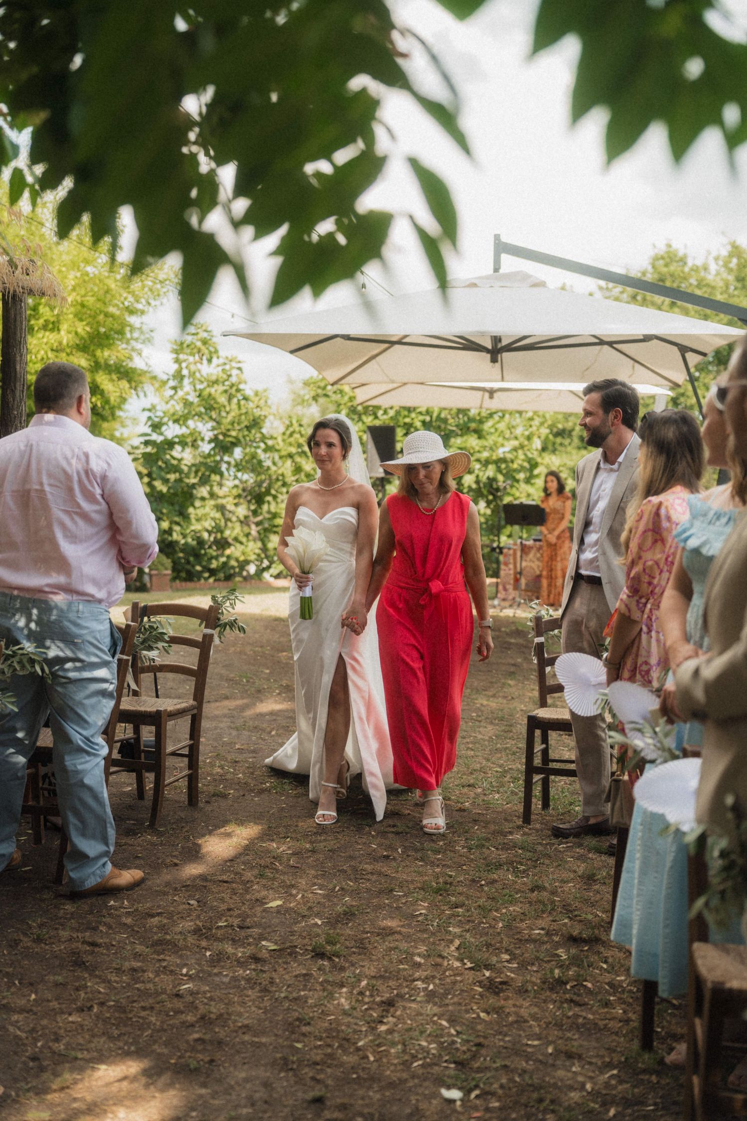 Bride in white gown walks down outdoor aisle with woman in red dress at garden wedding ceremony.