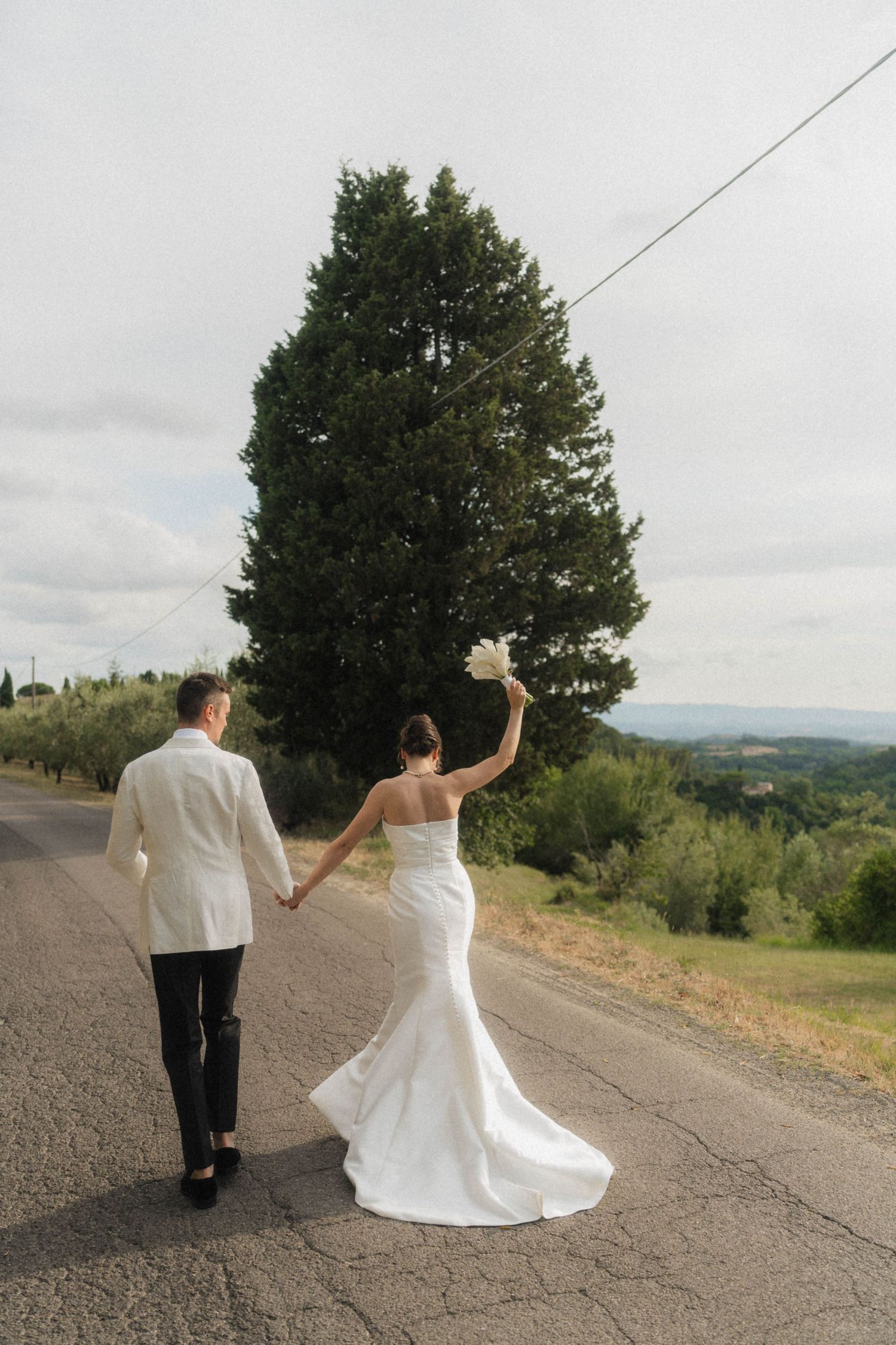 Bride and groom walking hand in hand down a road in Tuscany, bride raising white bouquet with cypress tree behind.