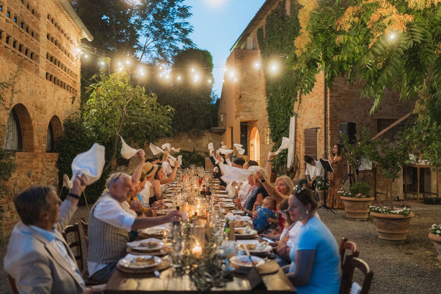 Wedding guests celebrate at long candlelit outdoor dinner table under string lights at Tuscan villa.
