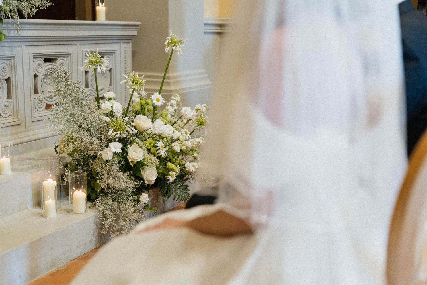 Bride in white gown and veil seated near elegant white floral arrangement with candles at church altar.