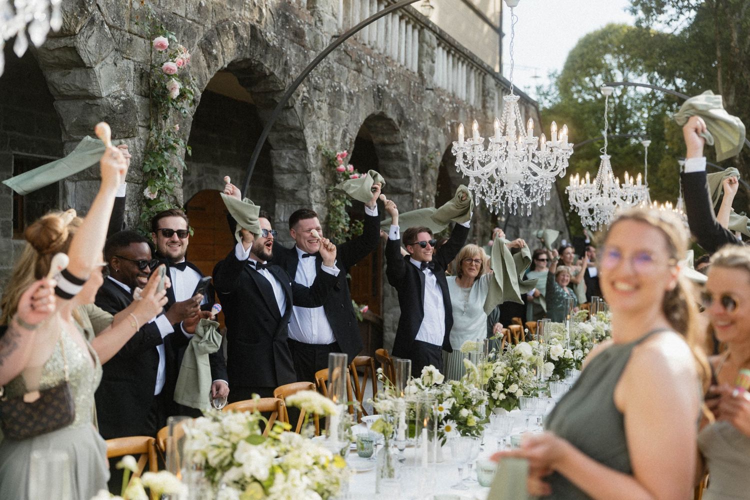 Wedding guests in formal attire raise glasses in toast at long outdoor reception table with chandeliers.