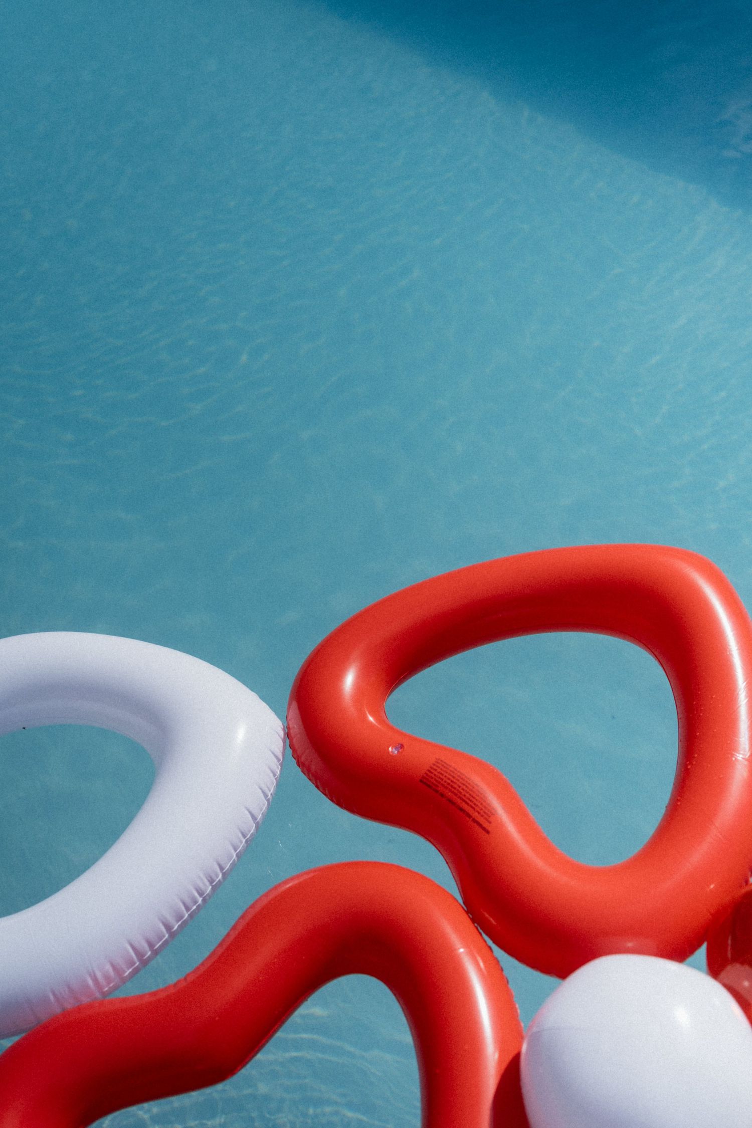 Red and white inflatable pool floats shaped like abstract curves floating in a bright blue swimming pool.