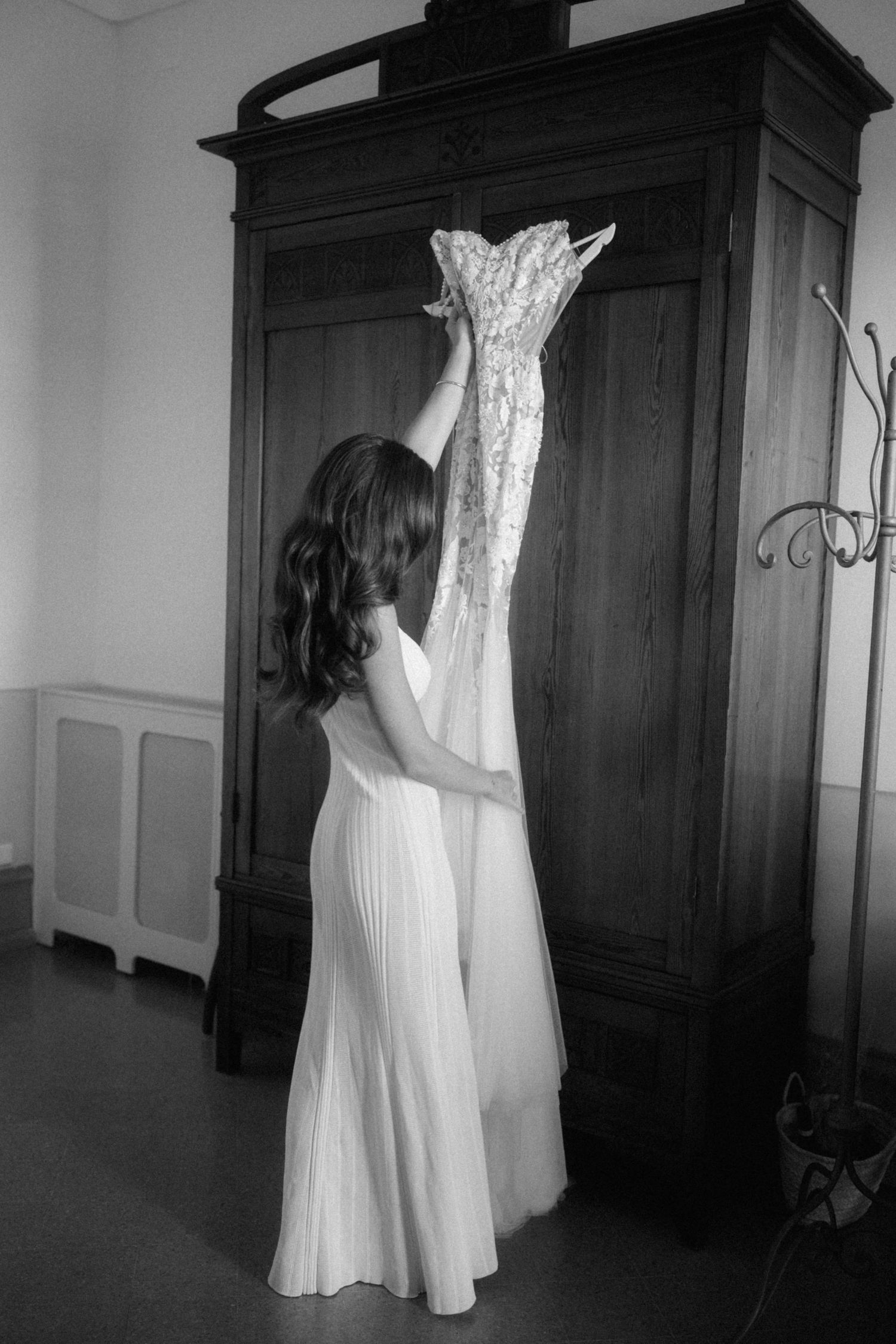 Bride in white dress reaching up to retrieve her lace wedding gown from a wooden armoire.