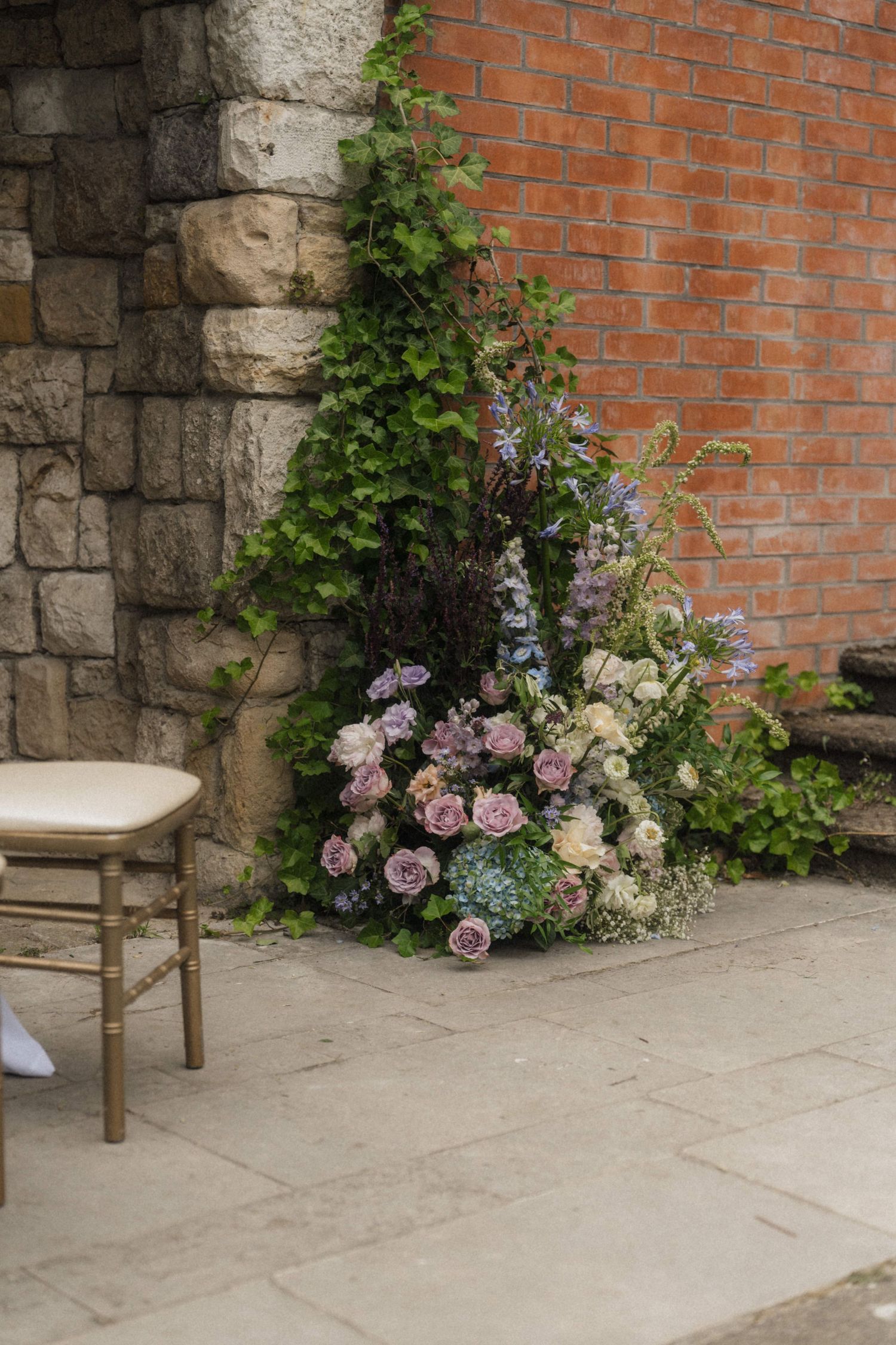 Elegant wedding floral arrangement of purple and white roses against a rustic brick wall with ivy, beside a gold chiavari chair.