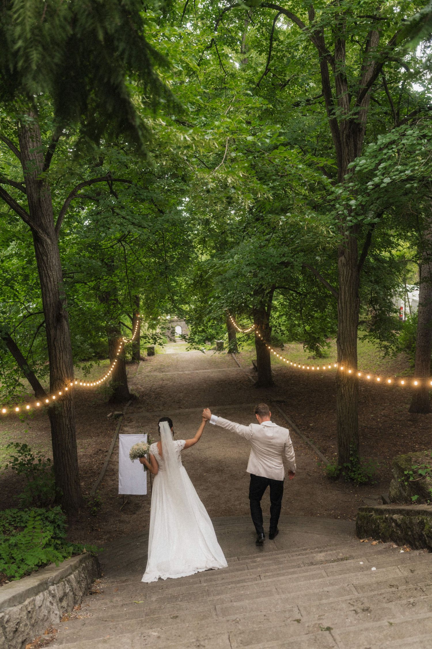 Bride and groom hold hands walking up stone steps through a lush green tree-lined path with string lights.