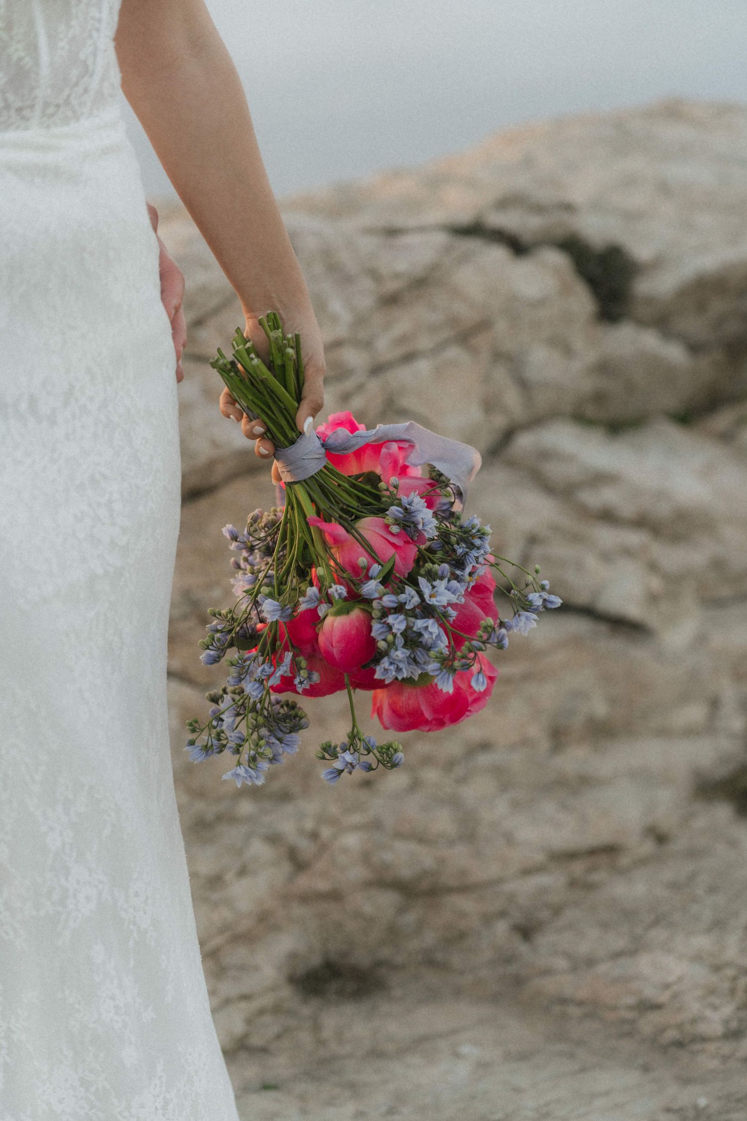 Bride in white lace gown holding pink peony and purple wildflower bouquet near rocky coast.