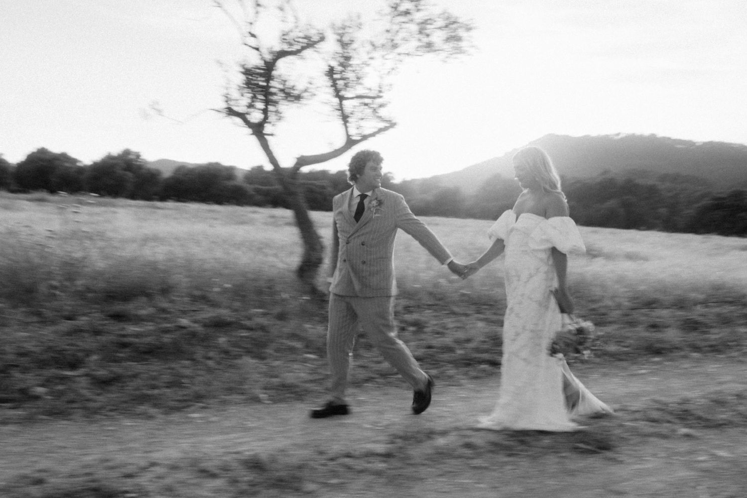 Black and white photo of joyful bride and groom running through a sunlit countryside field.