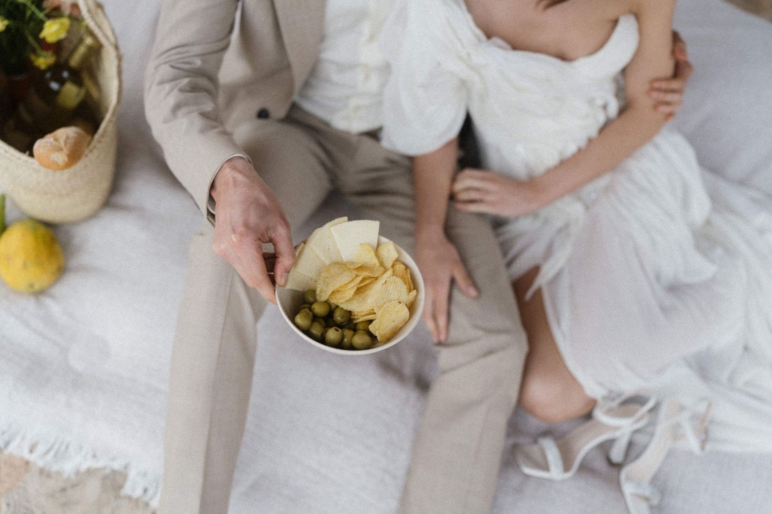 Groom in beige suit holds bowl of snacks beside bride in white wedding dress on white linen.