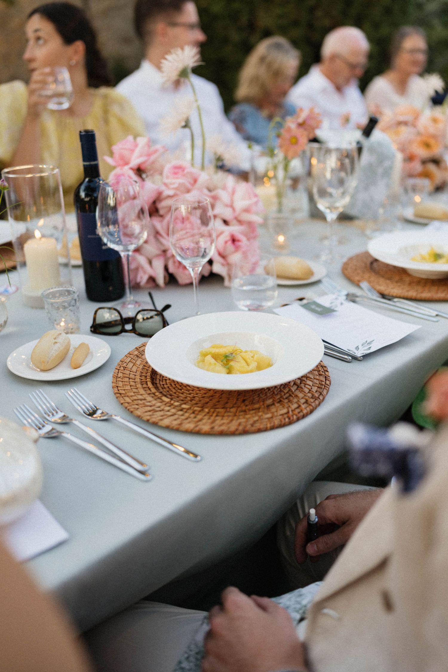 Elegant outdoor dinner party table set with pasta dishes, pink floral centerpieces, wine glasses, and candles.