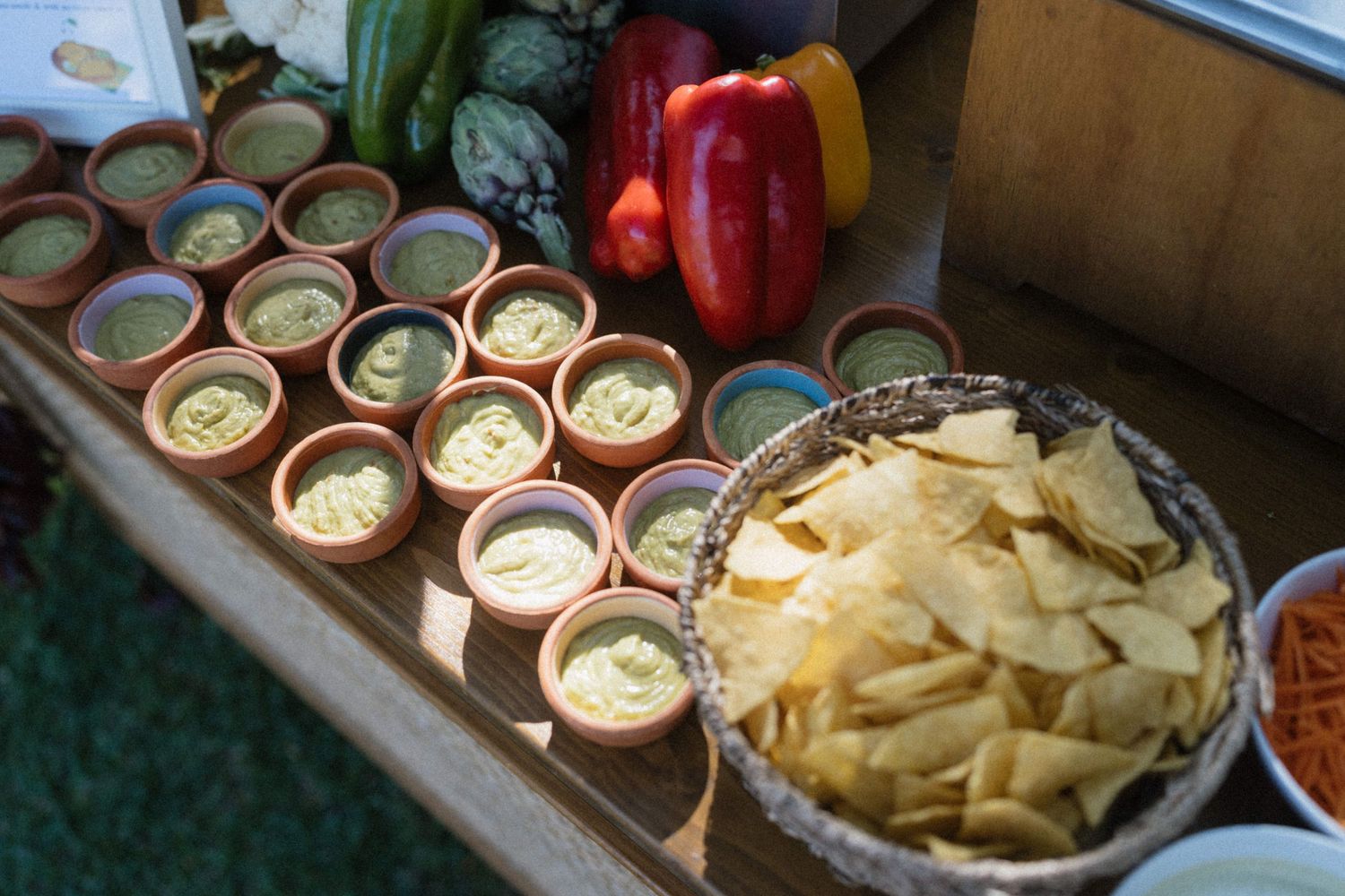 Clay cups filled with guacamole dip displayed on wooden board beside tortilla chips and fresh vegetables.