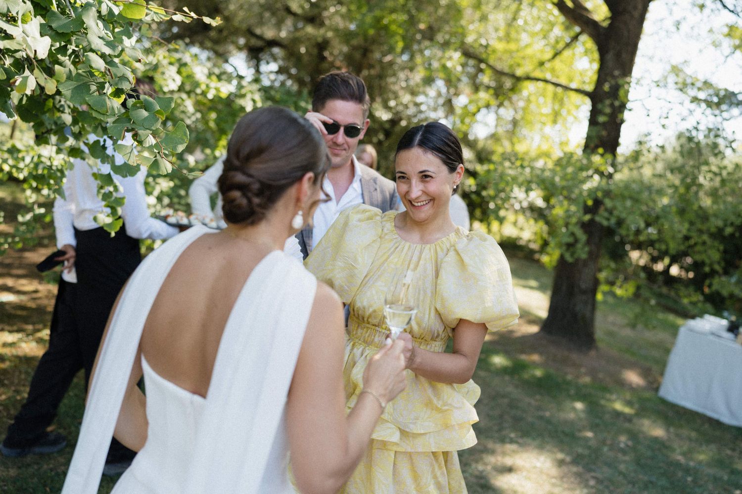 Bride in white gown toasts with guests in yellow dress and sunglasses at outdoor wedding reception.
