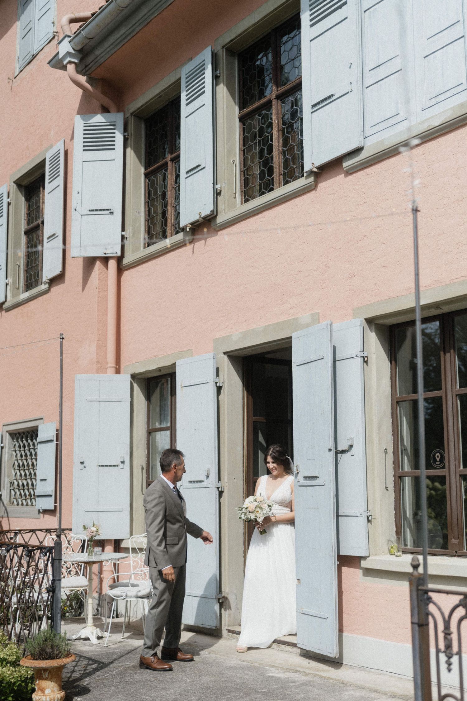 Bride in white gown holding bouquet greets groom outside pink building with blue shutters.