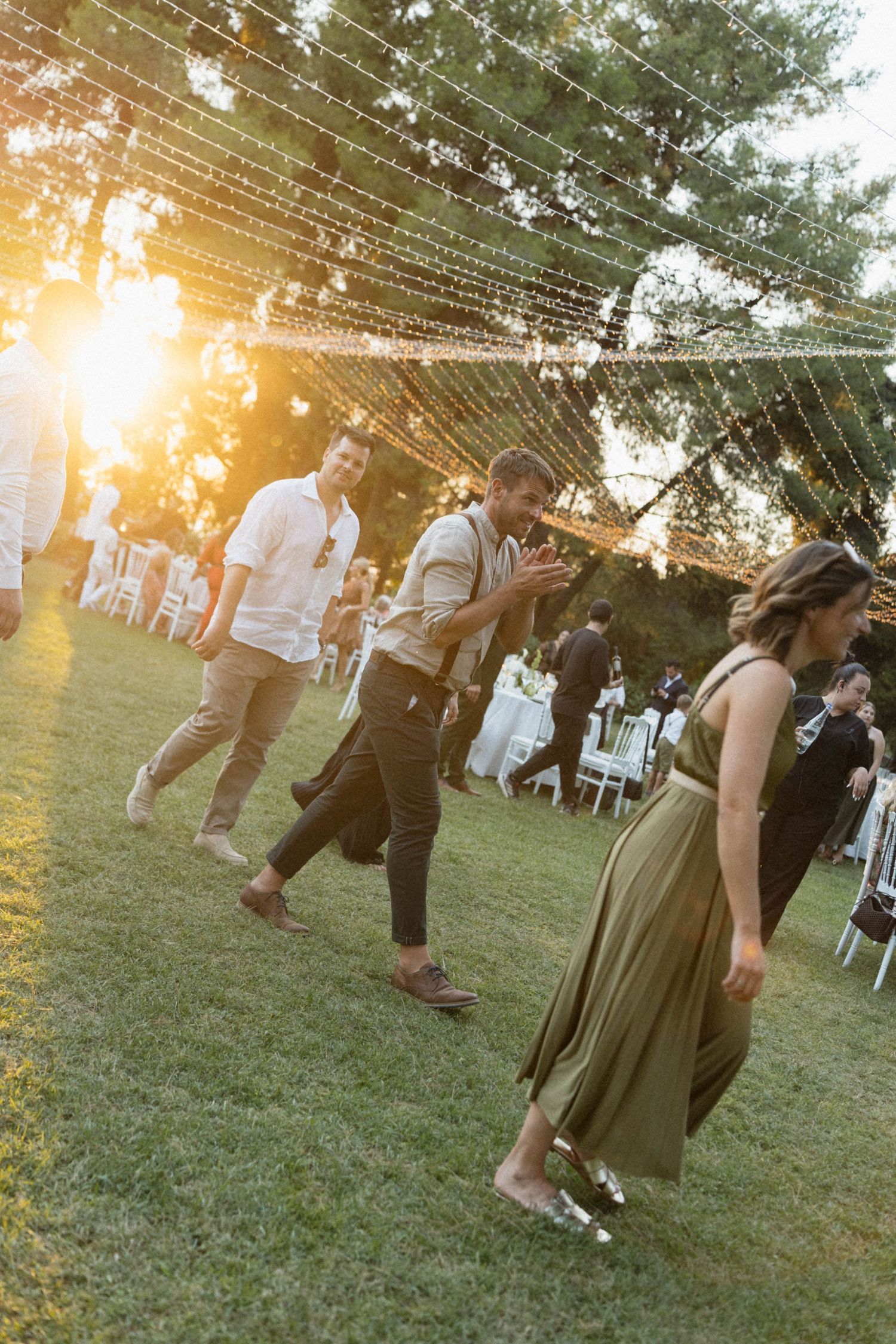 Guests walking on grass at an outdoor wedding reception at golden hour with string lights overhead.