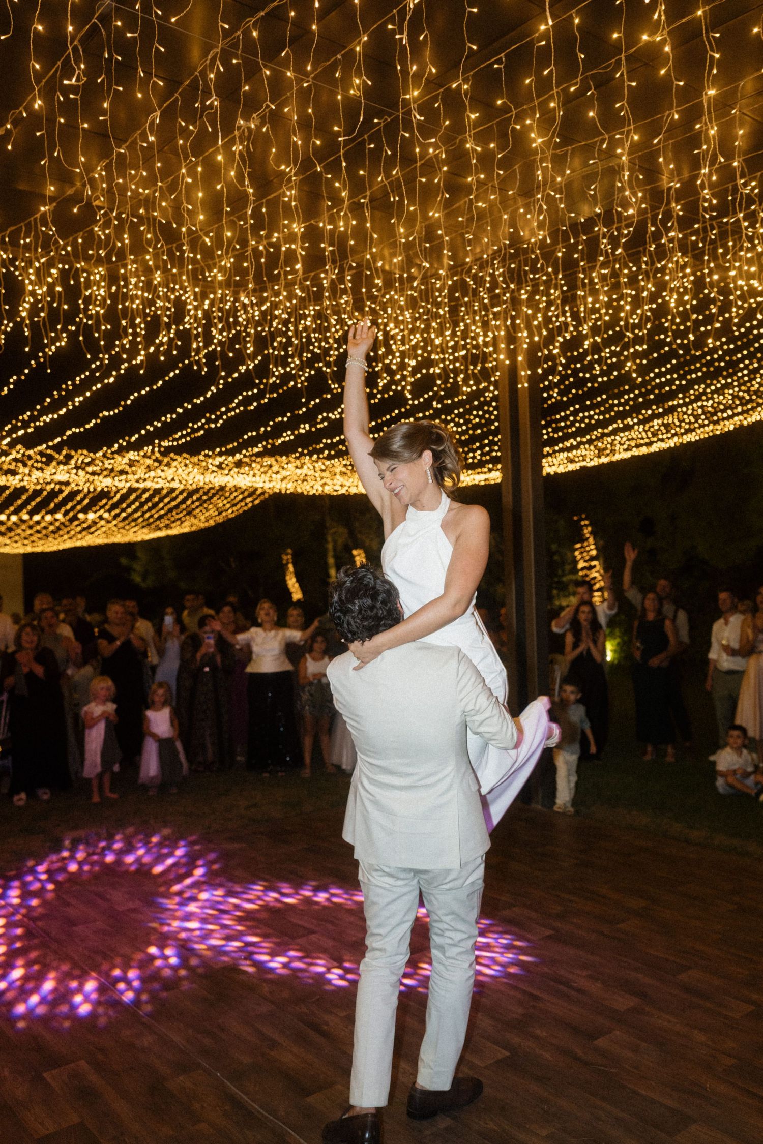 Groom lifts bride on wedding dance floor under sparkling fairy lights with guests watching in background.