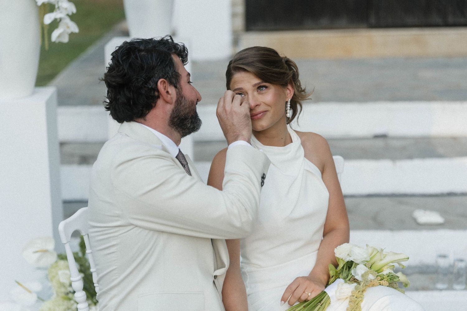 Groom gently wipes tears from bride's cheek during emotional outdoor wedding ceremony, both dressed in white.