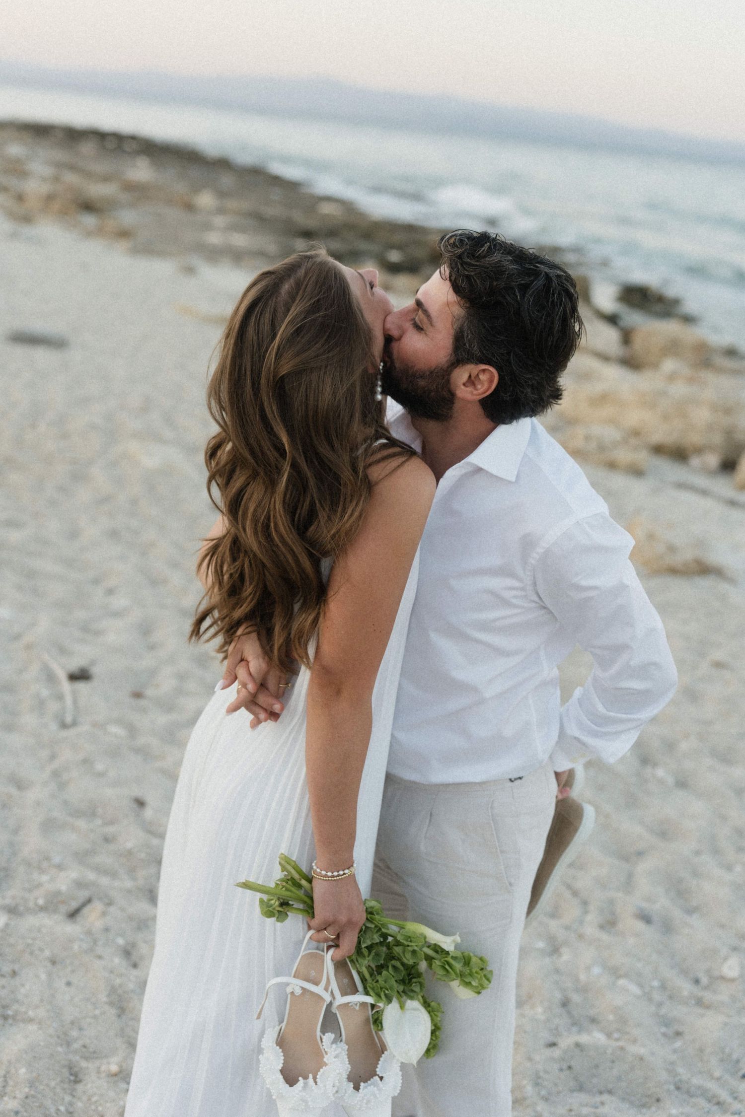 A couple shares a romantic kiss on a sandy beach, the bride in a white backless dress holding a green bouquet.