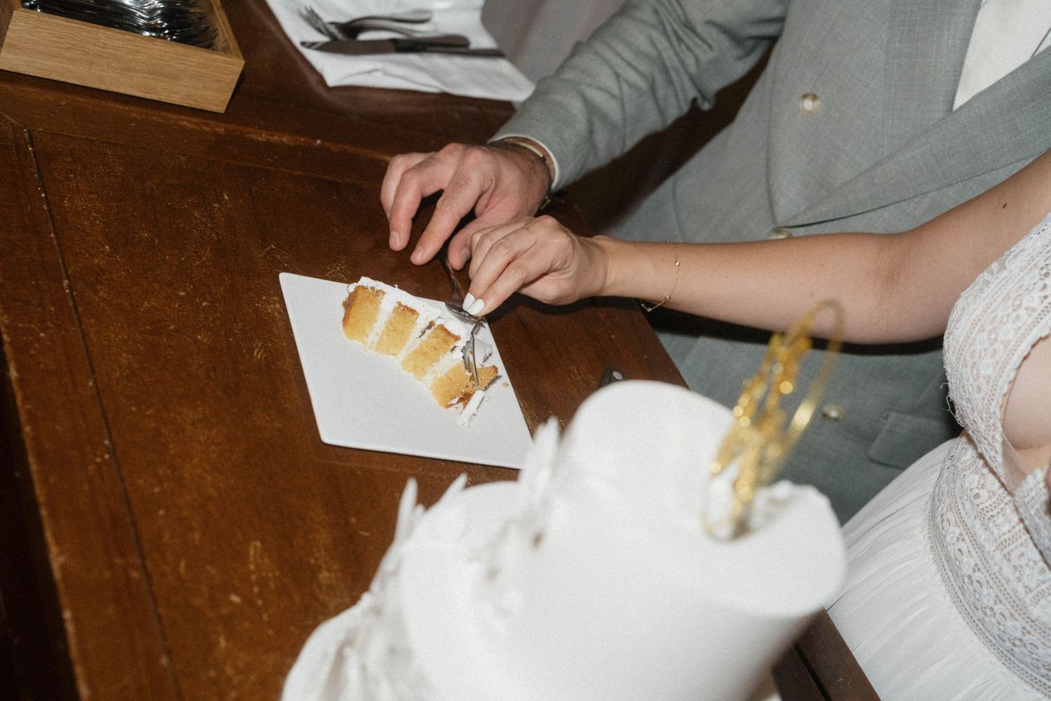 Bride and groom cutting a white wedding cake with gold decorations at a wooden table.