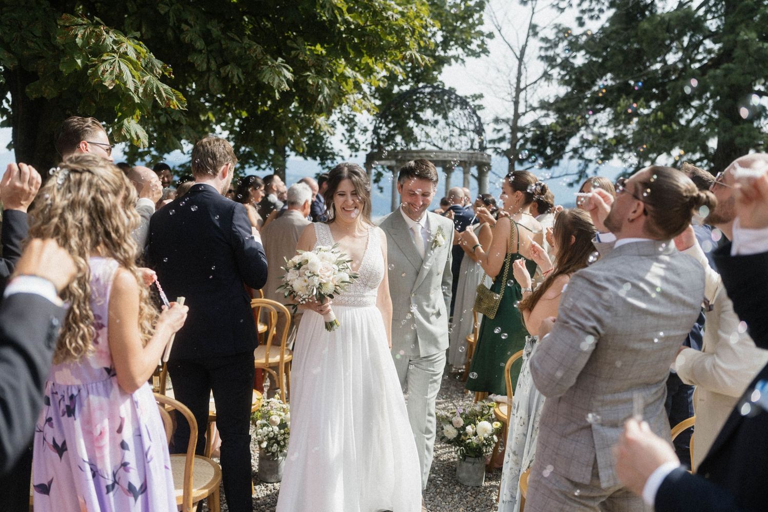 Bride and groom walk down the aisle as guests celebrate with confetti at an outdoor lakeside wedding.