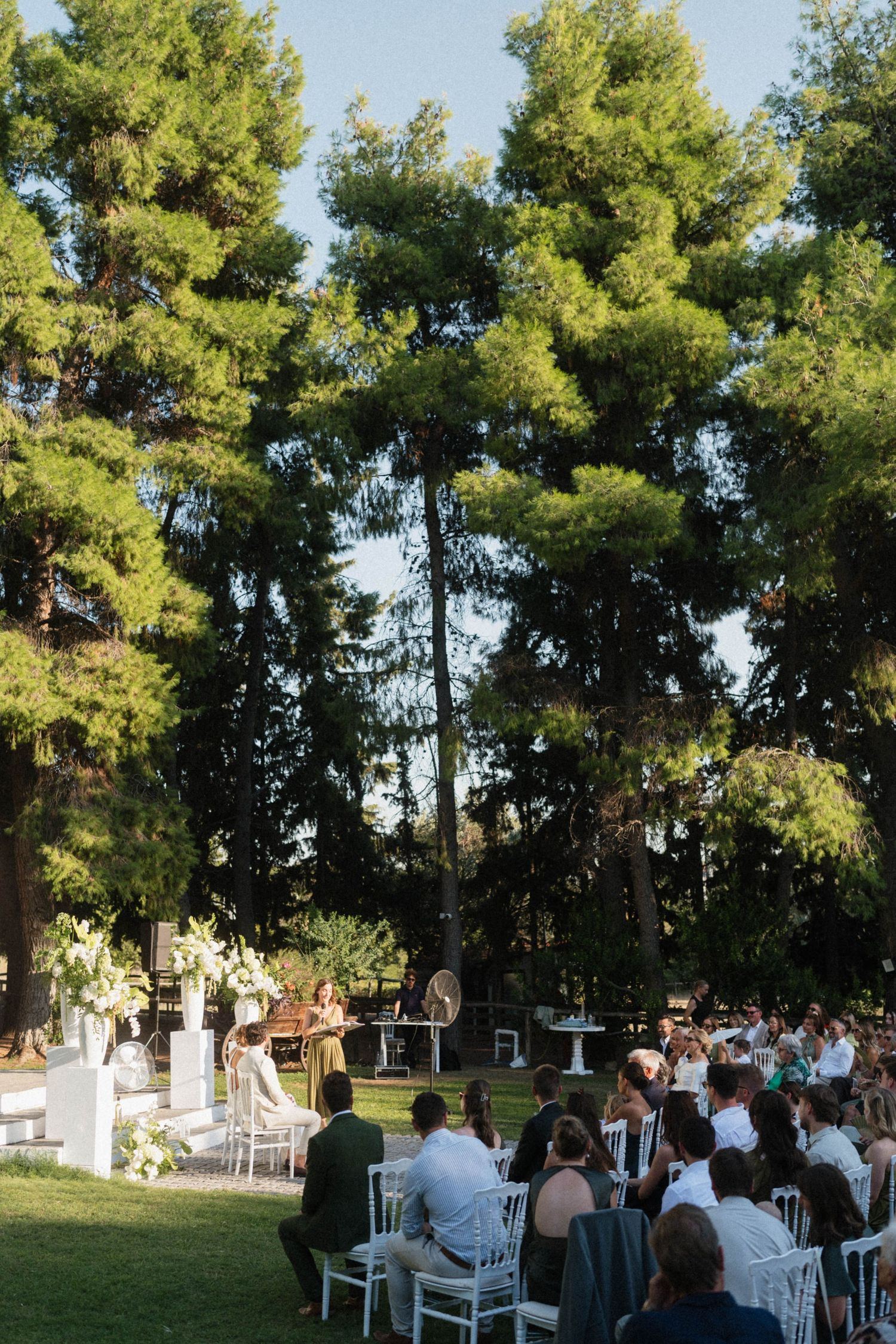 Guests seated in white chairs at an outdoor wedding ceremony surrounded by towering green trees in a garden setting.